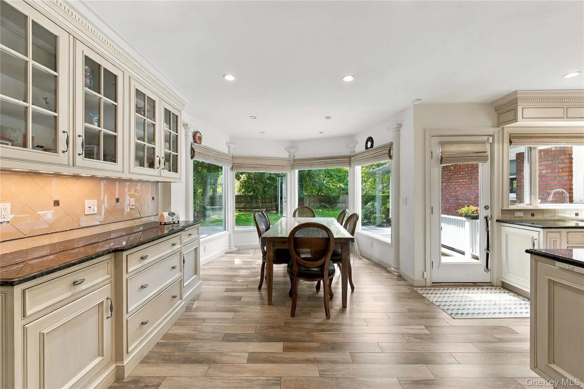 Kitchen featuring custom cabinetry with glass-front uppers, stone countertops, tiled backsplash, wood-finish flooring, and a bay window configuration Kitchen featuring custom cabinetry with glass-front uppers, stone countertops, tiled backsplash, wood-finish flooring, and a bay window configuration