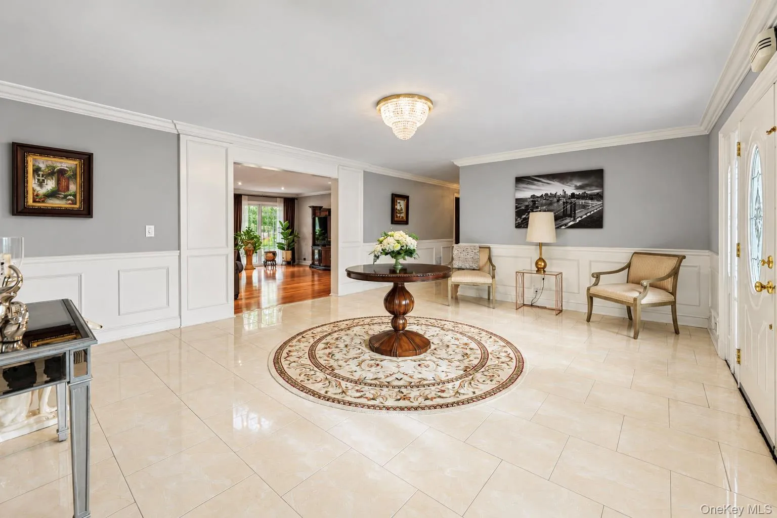 Foyer featuring large-format tile flooring, crisp wainscoting, crown molding, a decorative crystal-style ceiling fixture, and a white entry door with glass paneling Foyer featuring large-format tile flooring, crisp wainscoting, crown molding, a decorative crystal-style ceiling fixture, and a white entry door with glass paneling