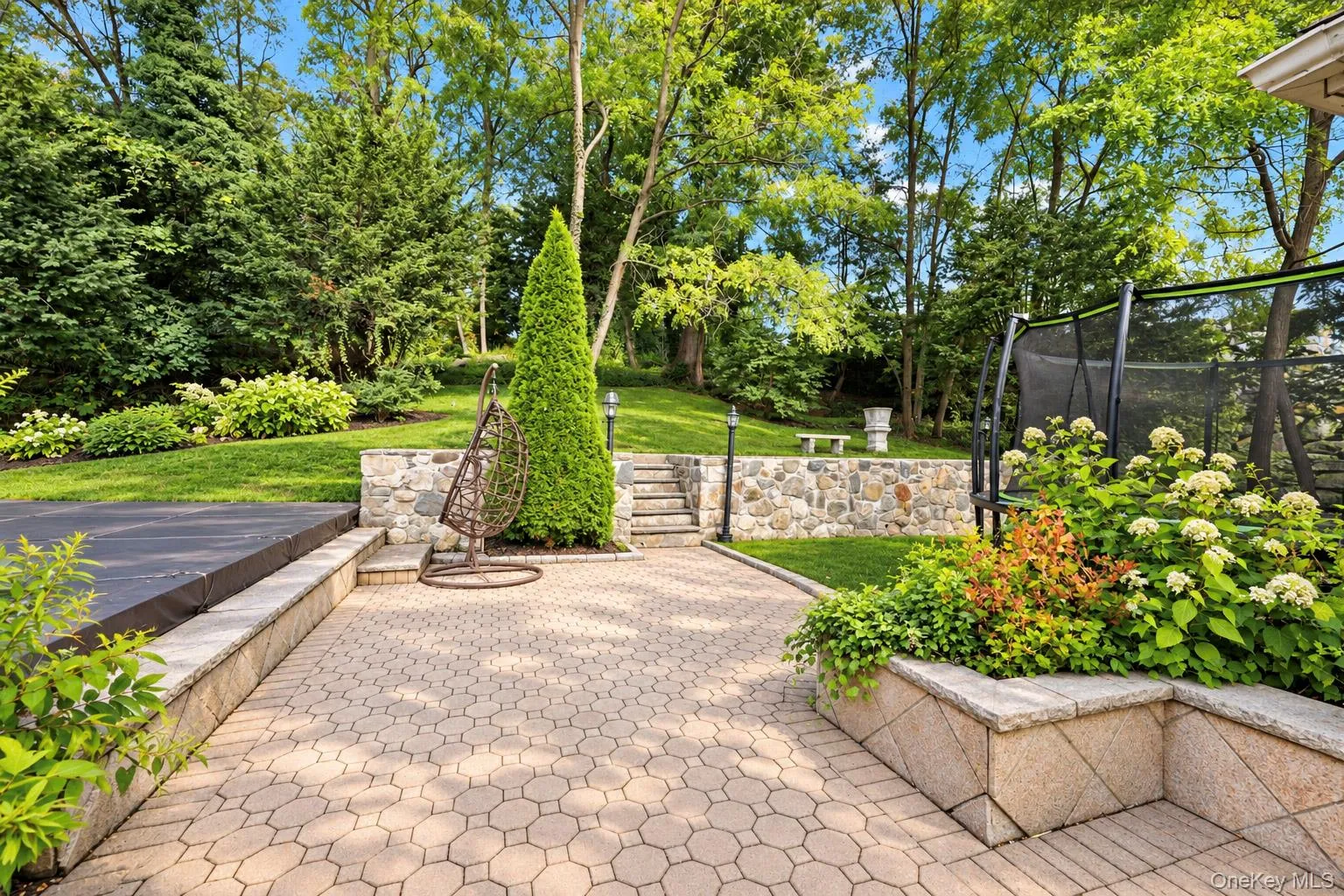 Paver patio featuring stone retaining walls, integrated stairs, tiered landscaping, and mature trees Paver patio featuring stone retaining walls, integrated stairs, tiered landscaping, and mature trees