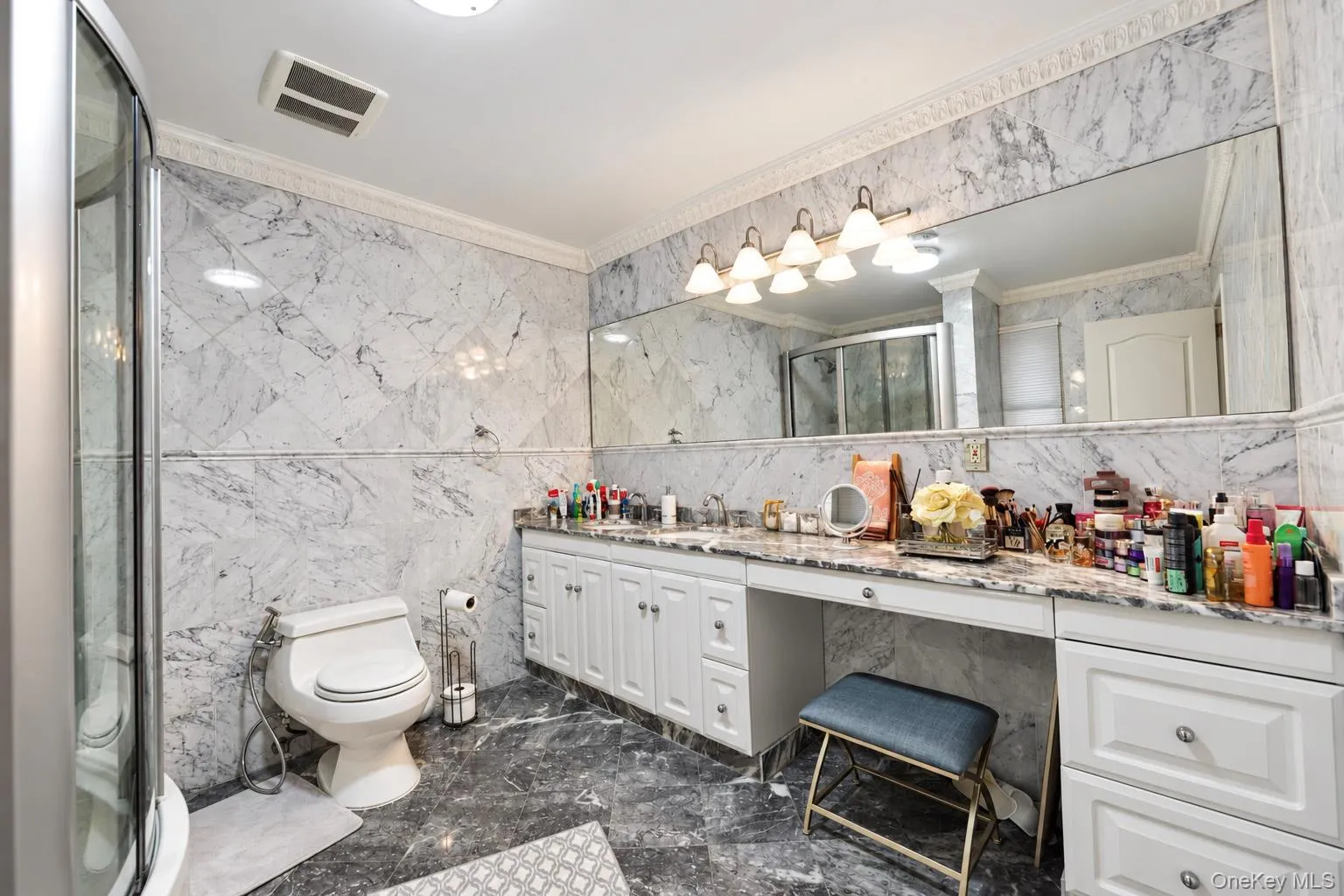 Full-bathroom featuring natural stone wall and floor tiling, expansive vanity with integrated stone countertop, crown molding, and multi-bulb wall-mounted lighting Full-bathroom featuring natural stone wall and floor tiling, expansive vanity with integrated stone countertop, crown molding, and multi-bulb wall-mounted lighting