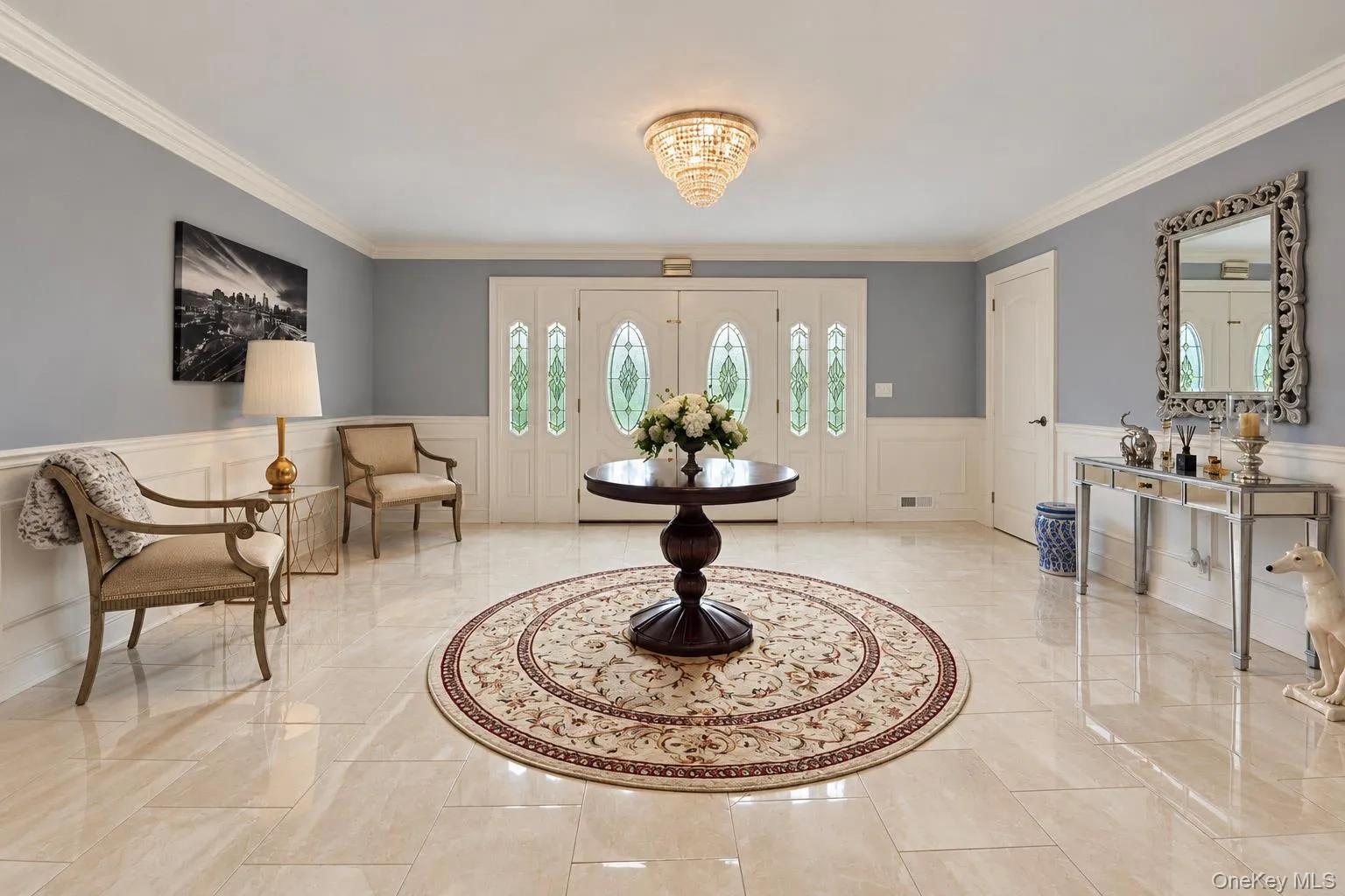 Foyer featuring large-format tile flooring, double doors with decorative glass inserts, crown molding, and wainscoting, accented by a tiered crystal-style flush-mount light fixture Foyer featuring large-format tile flooring, double doors with decorative glass inserts, crown molding, and wainscoting, accented by a tiered crystal-style flush-mount light fixture