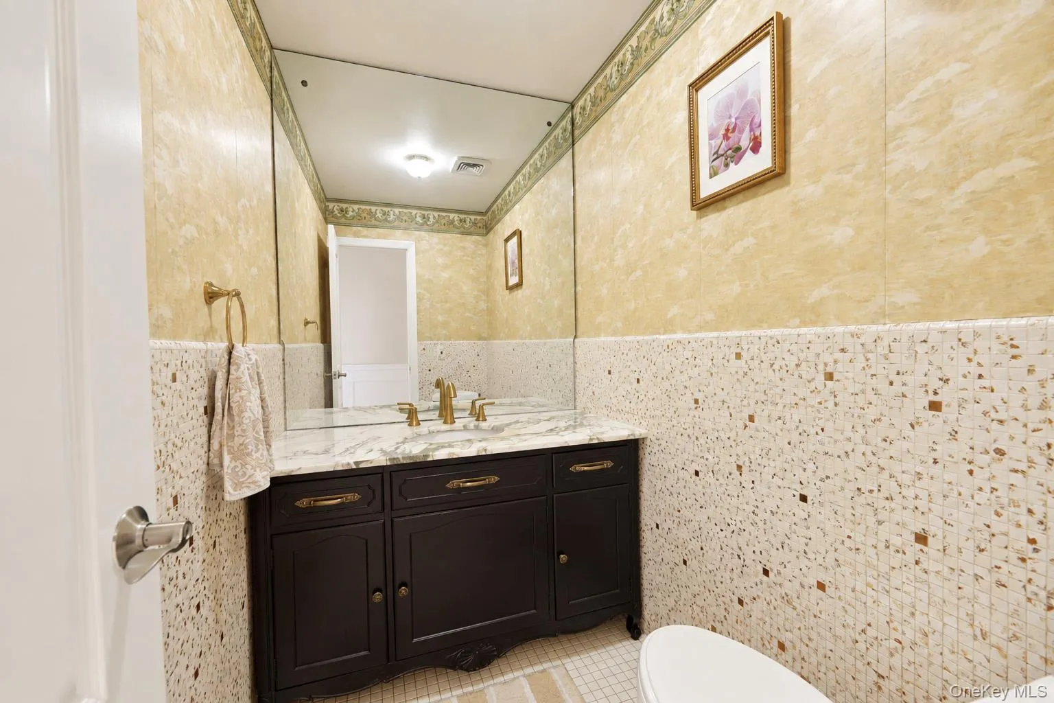 Dark-finish vanity featuring a stone-surface countertop, gold-toned faucet hardware, a wall-to-wall mirror, and textured tile wall accents Dark-finish vanity featuring a stone-surface countertop, gold-toned faucet hardware, a wall-to-wall mirror, and textured tile wall accents