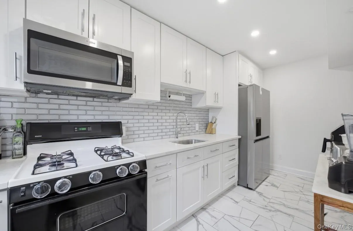 Kitchen featuring white shaker-style cabinetry, stainless steel appliances, a light-toned stone-finish countertop, a horizontal subway tile backsplash, and large-format tile flooring with a veined pattern Kitchen featuring white shaker-style cabinetry, stainless steel appliances, a light-toned stone-finish countertop, a horizontal subway tile backsplash, and large-format tile flooring with a veined pattern