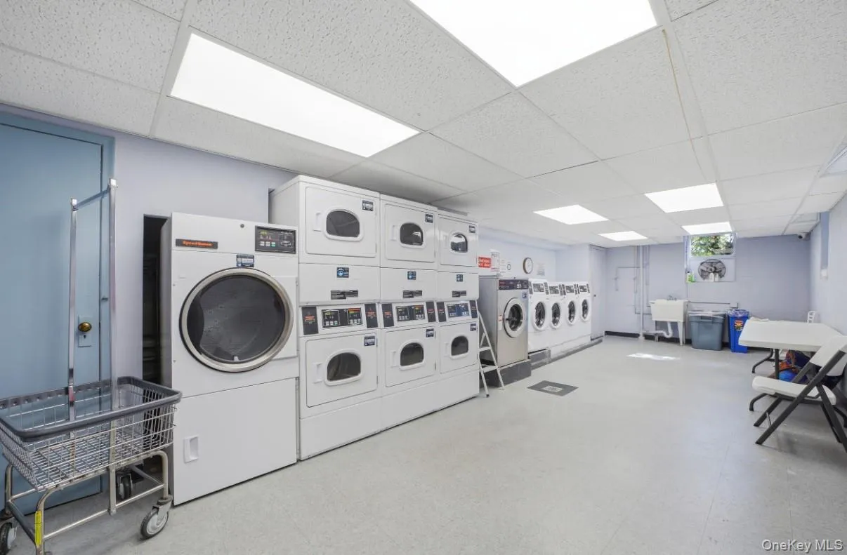 Laundry room featuring multiple stackable drying units, a standard washing machine, a utility sink, and drop ceiling panels with integrated lighting Laundry room featuring multiple stackable drying units, a standard washing machine, a utility sink, and drop ceiling panels with integrated lighting