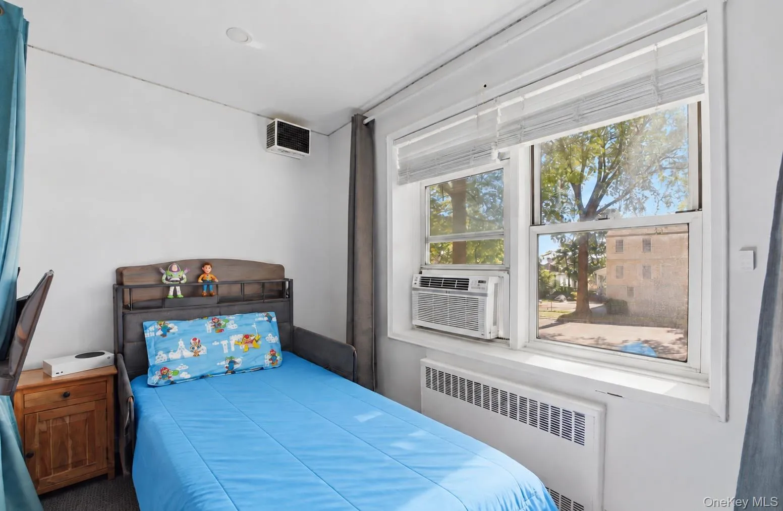 Bright room featuring double-pane windows with integrated blinds, a built-in window-mounted air conditioning unit, and a metal radiator cover Bright room featuring double-pane windows with integrated blinds, a built-in window-mounted air conditioning unit, and a metal radiator cover