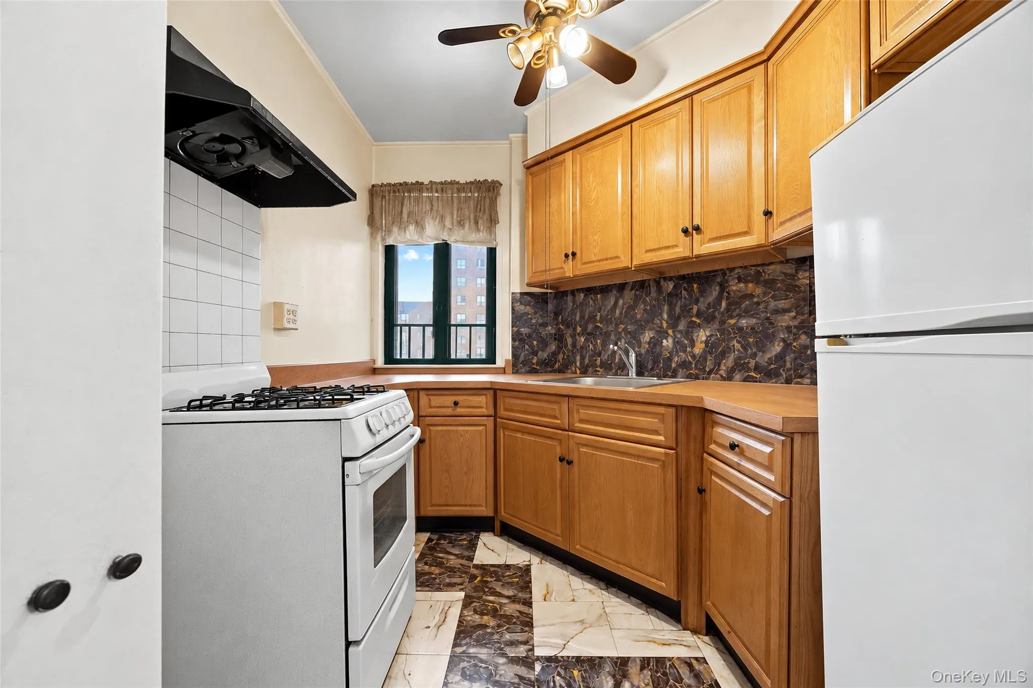 Kitchen featuring natural wood cabinetry, stone-patterned backsplash, geometric tiled flooring, gas range with an overhead vent hood, and a ceiling-mounted light fixture Kitchen featuring natural wood cabinetry, stone-patterned backsplash, geometric tiled flooring, gas range with an overhead vent hood, and a ceiling-mounted light fixture
