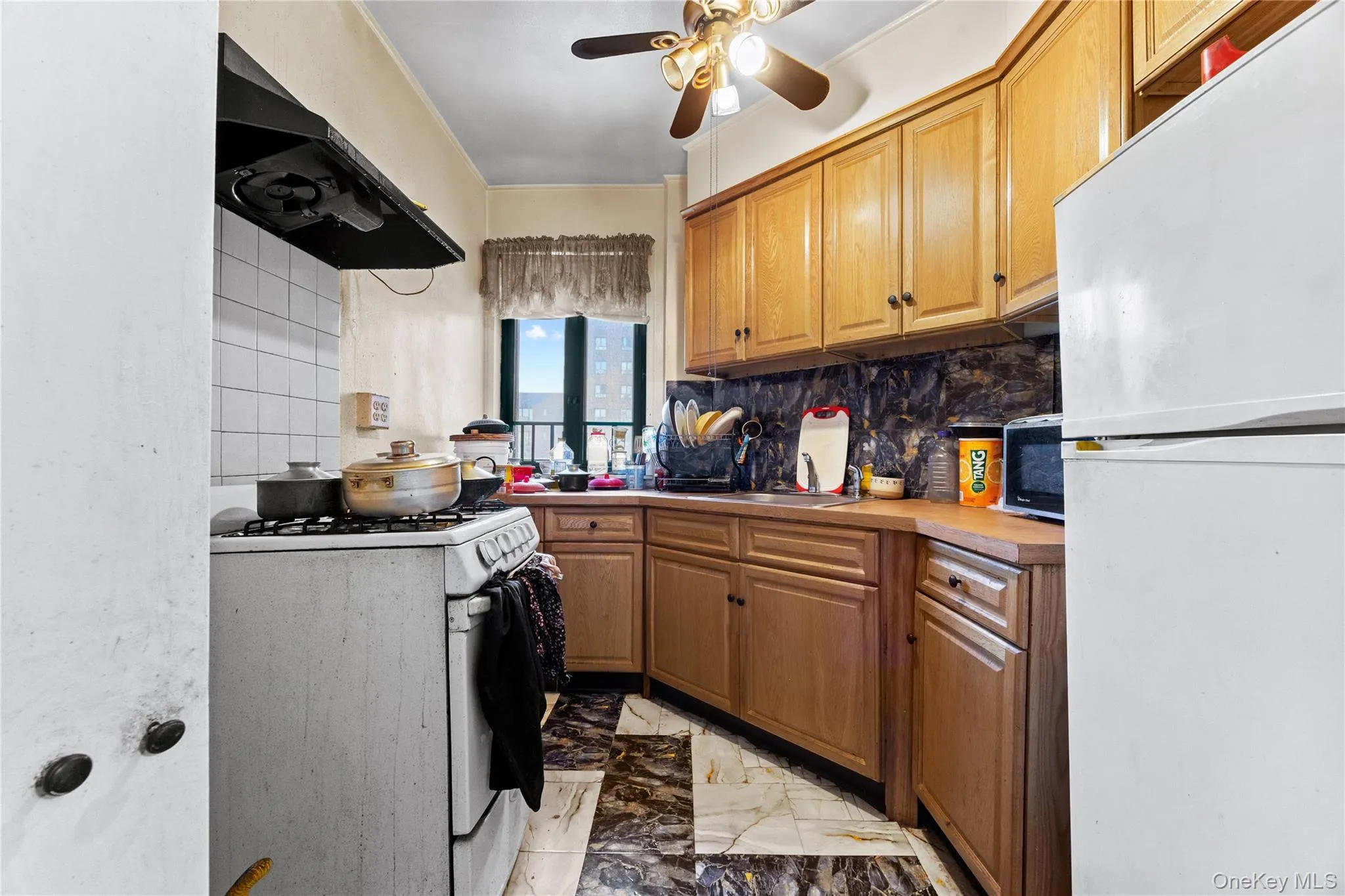 Kitchen featuring natural wood cabinetry, stone-patterned backsplash, geometric stone-tile flooring, and a ceiling-mounted light fixture with integrated fan Kitchen featuring natural wood cabinetry, stone-patterned backsplash, geometric stone-tile flooring, and a ceiling-mounted light fixture with integrated fan