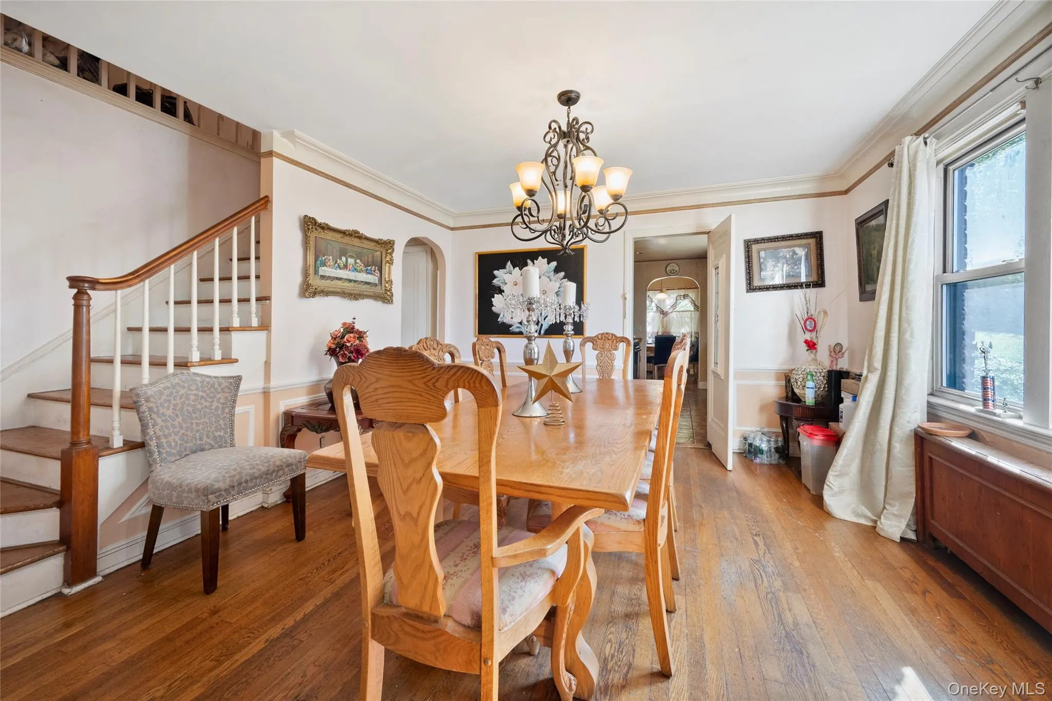 Dining room featuring wood-finish flooring, wainscoting, an ornate iron chandelier, and a staircase with a stained wood handrail Dining room featuring wood-finish flooring, wainscoting, an ornate iron chandelier, and a staircase with a stained wood handrail