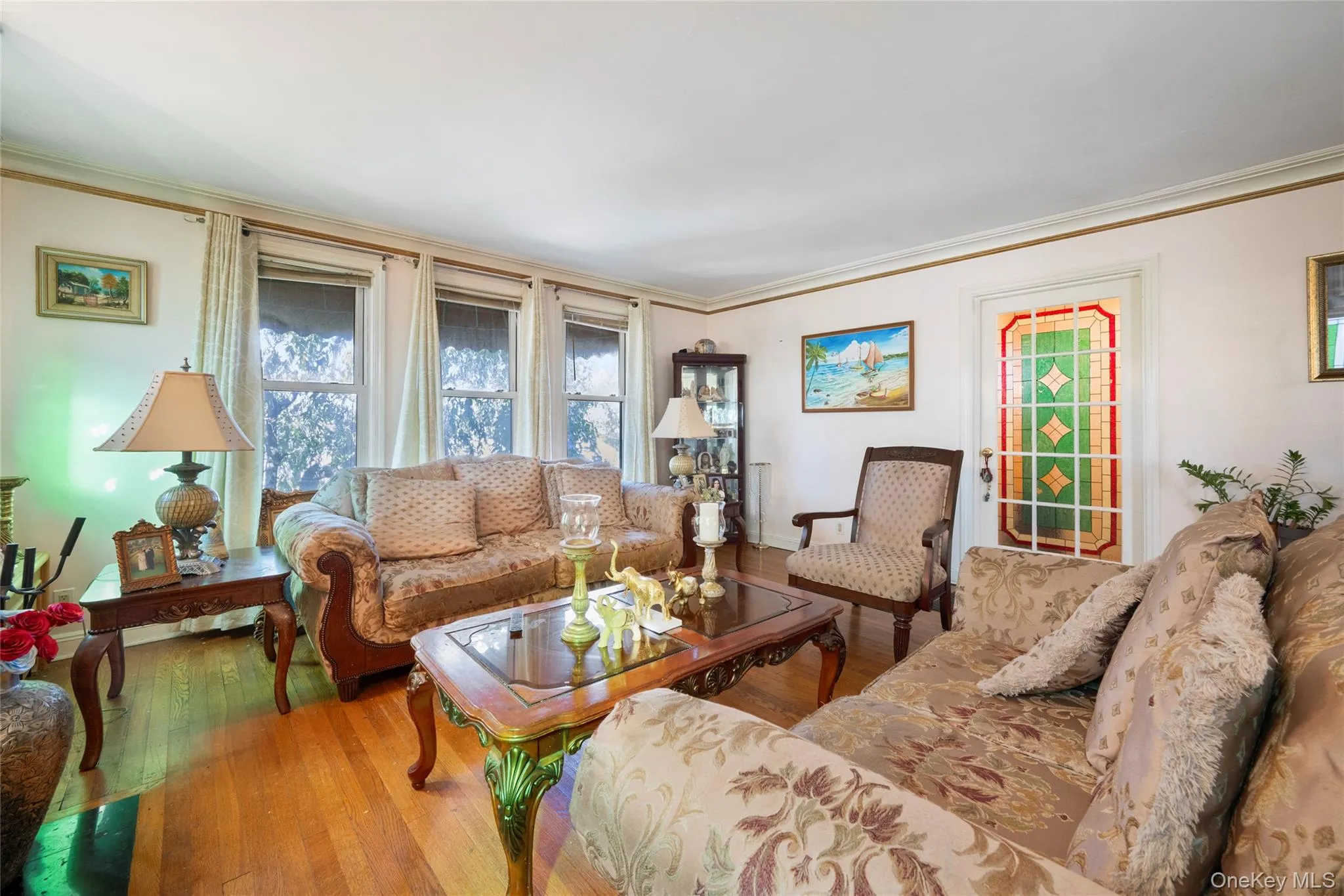 Living area featuring wood-finish flooring, crown molding, three large windows with neutral drapery, and a classic door with decorative stained-glass inlay Living area featuring wood-finish flooring, crown molding, three large windows with neutral drapery, and a classic door with decorative stained-glass inlay