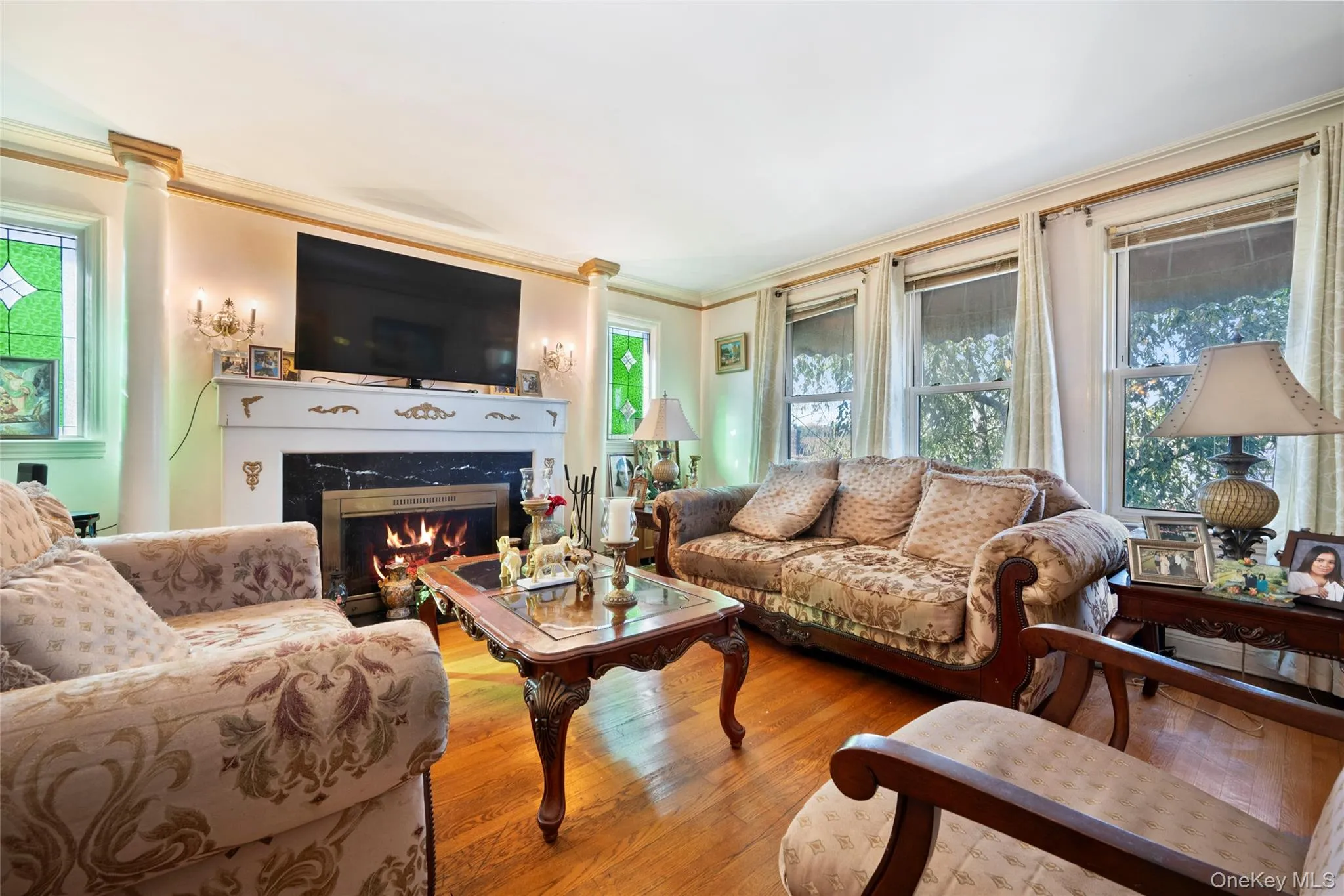 Living room featuring wood-finish flooring, a fireplace with a decorative mantel and stone surround, built-in column detailing, crown molding, and stained-glass accent windows Living room featuring wood-finish flooring, a fireplace with a decorative mantel and stone surround, built-in column detailing, crown molding, and stained-glass accent windows
