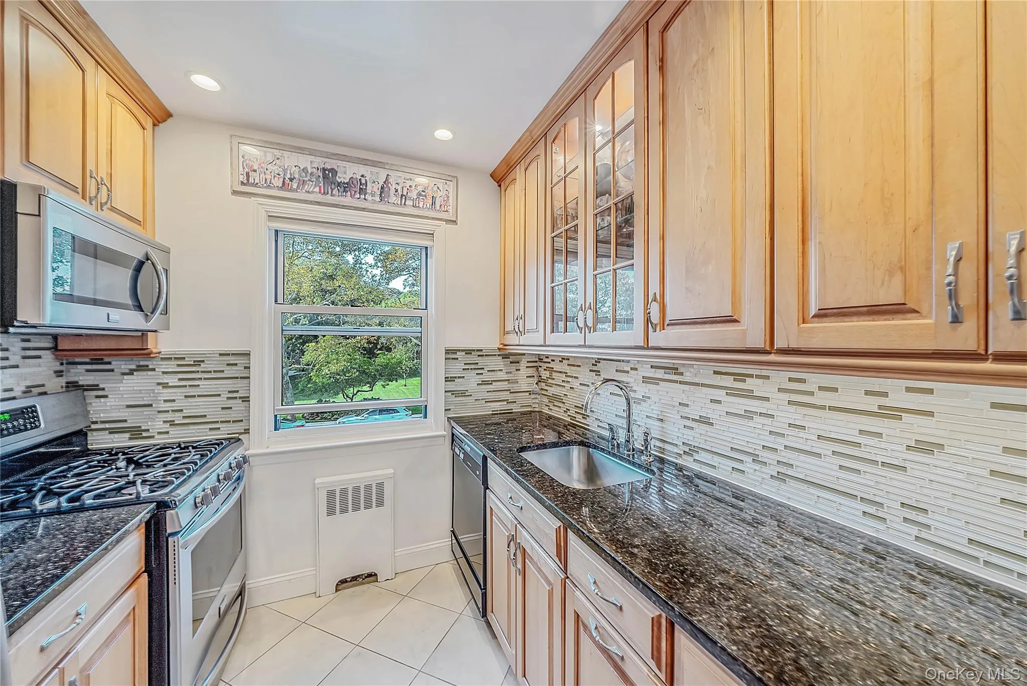 Kitchen featuring light wood cabinetry with glass-panel uppers, dark stone-finish countertops, multi-tonal linear tile backsplash, stainless steel appliances, and tile flooring Kitchen featuring light wood cabinetry with glass-panel uppers, dark stone-finish countertops, multi-tonal linear tile backsplash, stainless steel appliances, and tile flooring