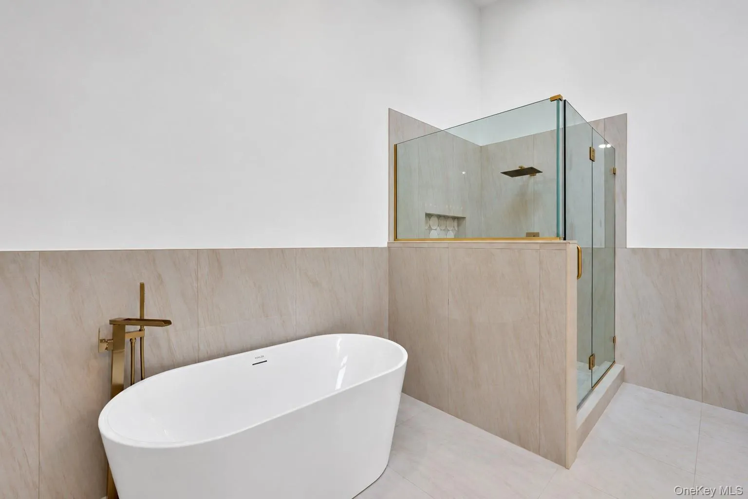 Freestanding white soaking tub with gold-tone floor-mounted faucet, walk-in glass shower enclosure, gold-tone shower fixtures, and neutral large-format tile wall cladding Freestanding white soaking tub with gold-tone floor-mounted faucet, walk-in glass shower enclosure, gold-tone shower fixtures, and neutral large-format tile wall cladding