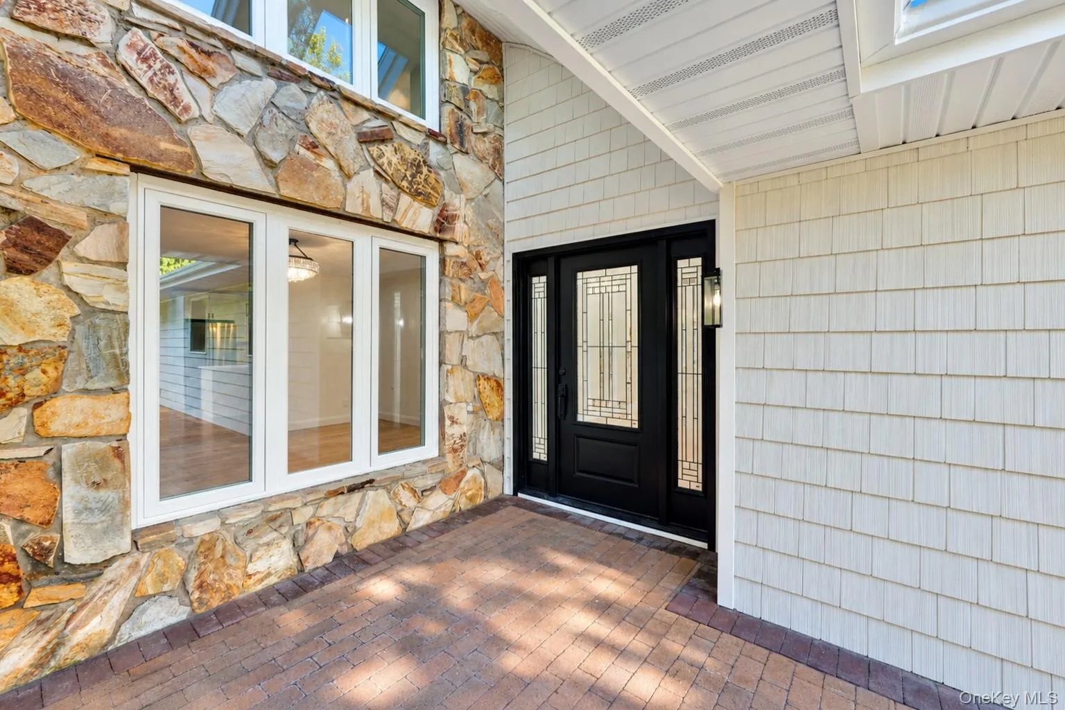Exterior entrance featuring multi-hued natural stone masonry, white shake siding, brick paver porch, and a black front door with decorative glass panels and sidelights Exterior entrance featuring multi-hued natural stone masonry, white shake siding, brick paver porch, and a black front door with decorative glass panels and sidelights