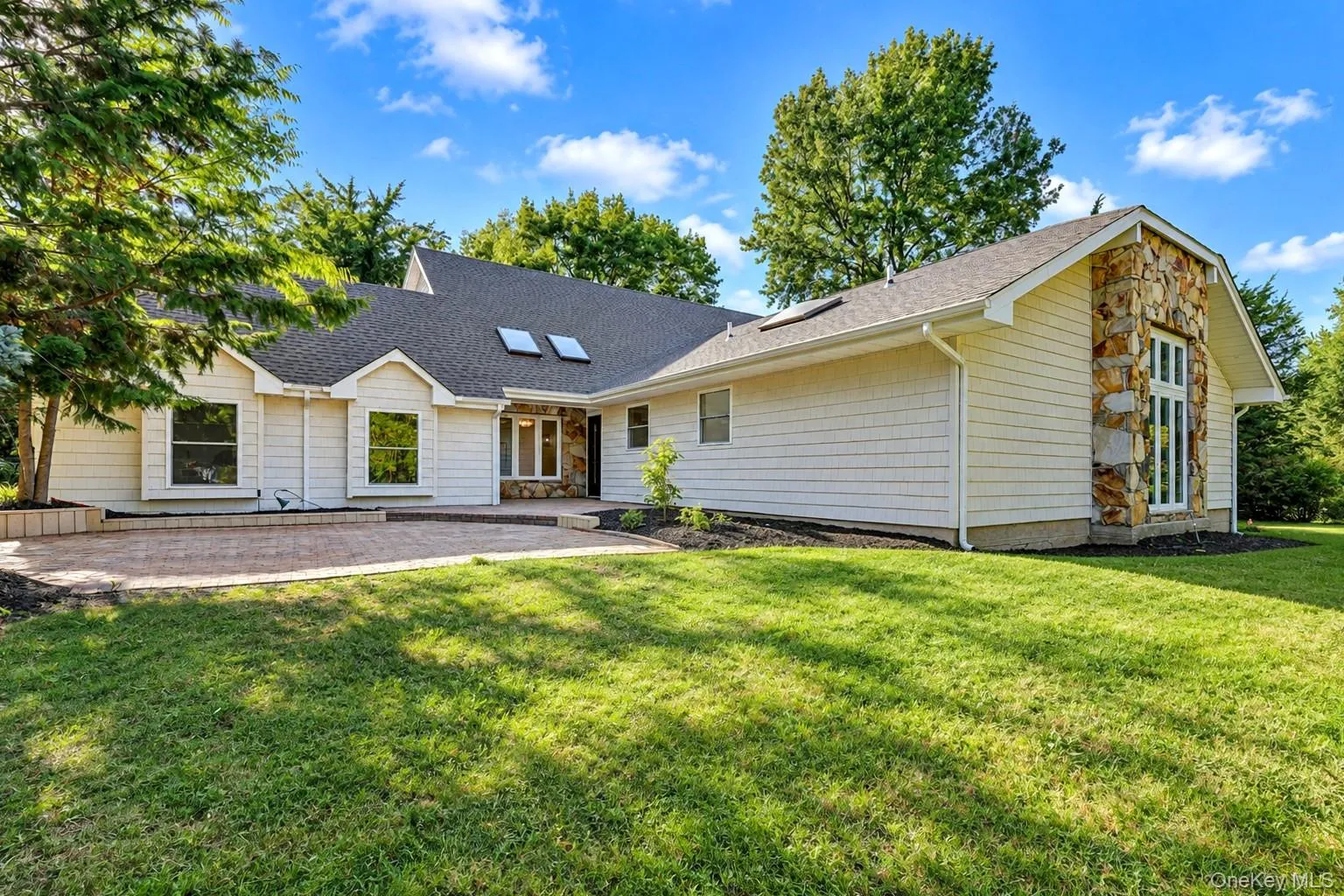 Exterior featuring light-toned shingle siding, natural stone accents, a brick paver patio, roof skylights, and a manicured lawn Exterior featuring light-toned shingle siding, natural stone accents, a brick paver patio, roof skylights, and a manicured lawn