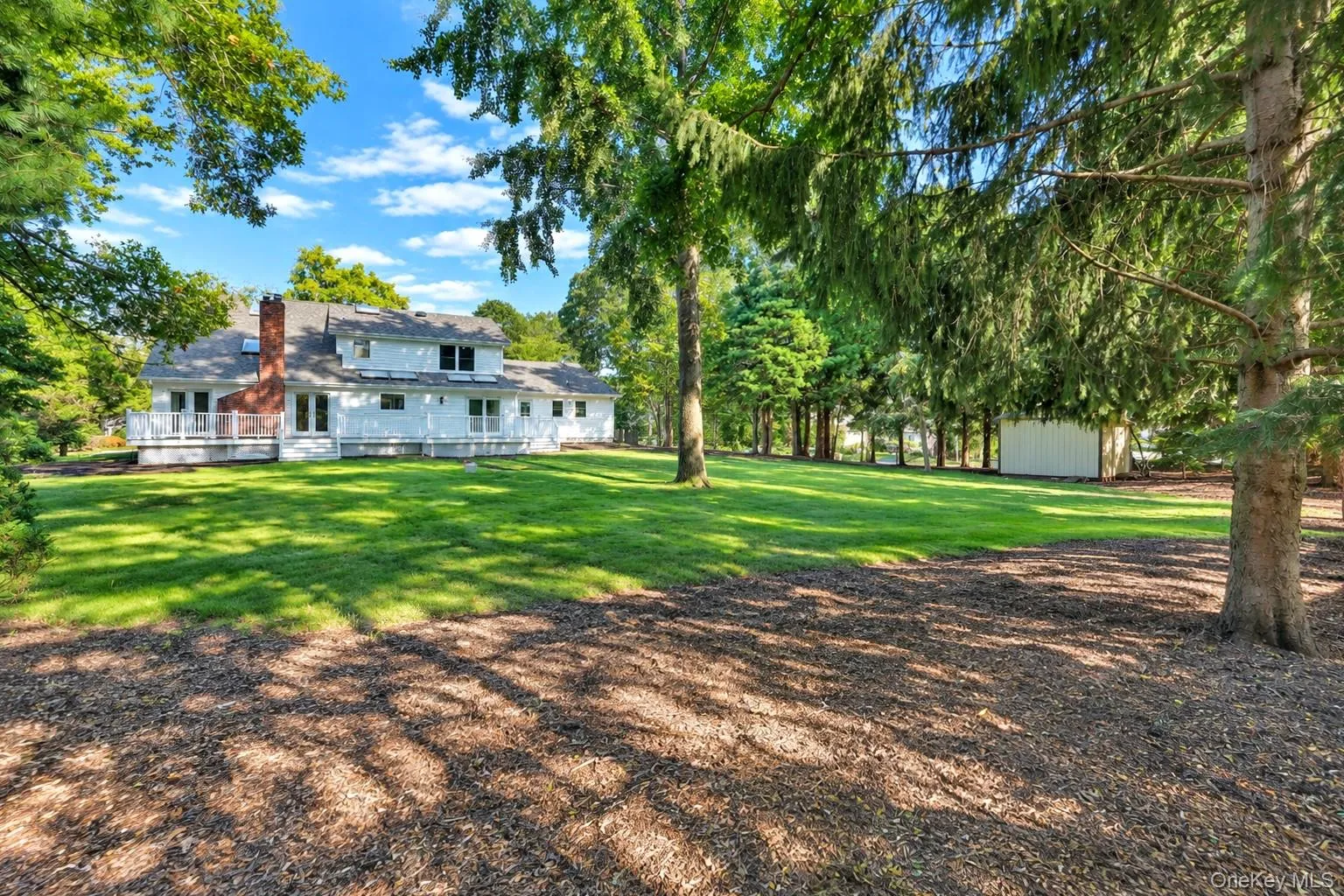 Expansive backyard featuring a manicured lawn, mature trees, a detached storage structure, and a raised wooden deck with white railing Expansive backyard featuring a manicured lawn, mature trees, a detached storage structure, and a raised wooden deck with white railing