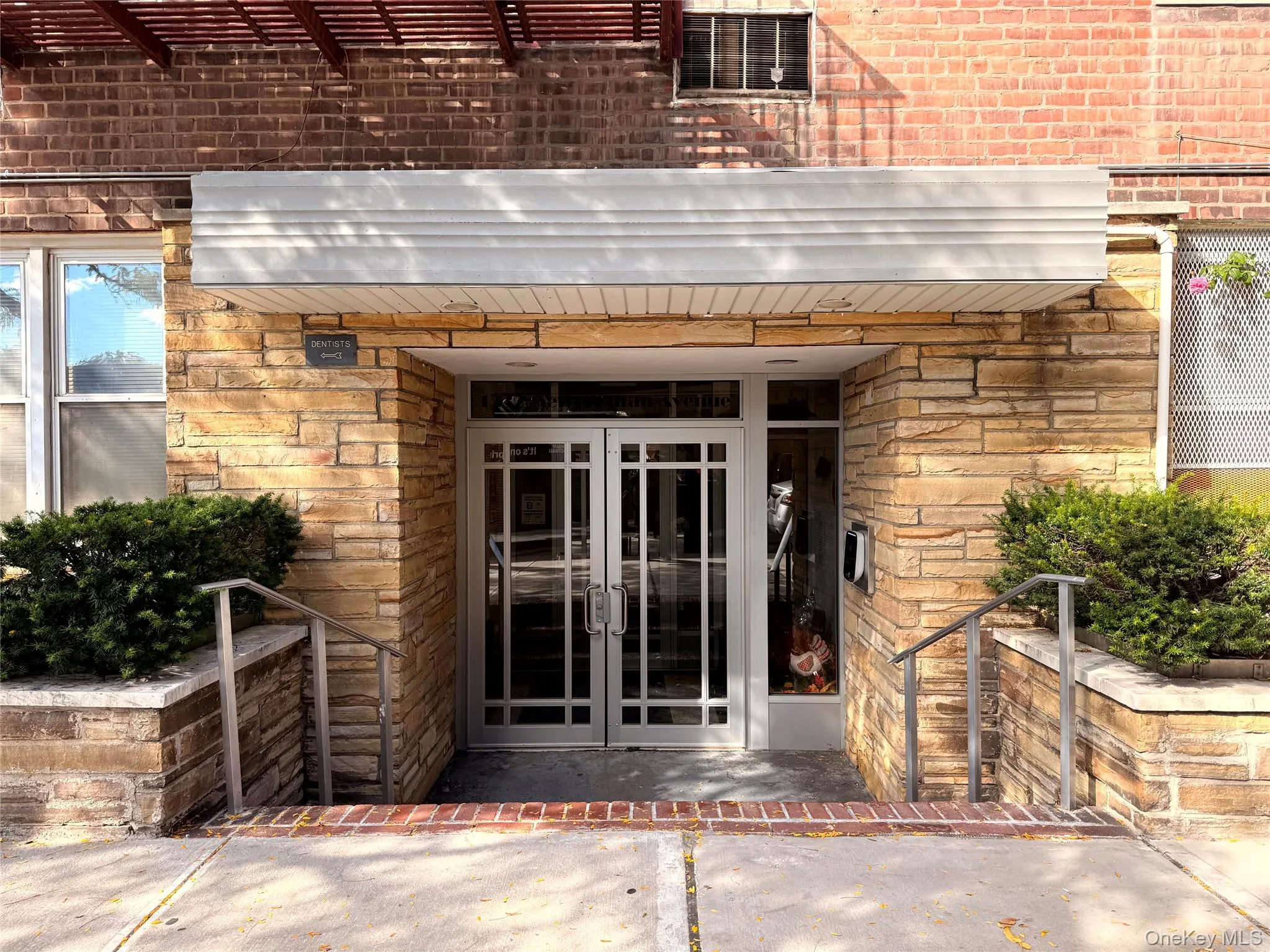 Building entrance featuring a textured stone facade, double glass entry doors with grid detailing, a prominent horizontal overhead canopy, and stainless steel handrails framing concrete steps Building entrance featuring a textured stone facade, double glass entry doors with grid detailing, a prominent horizontal overhead canopy, and stainless steel handrails framing concrete steps