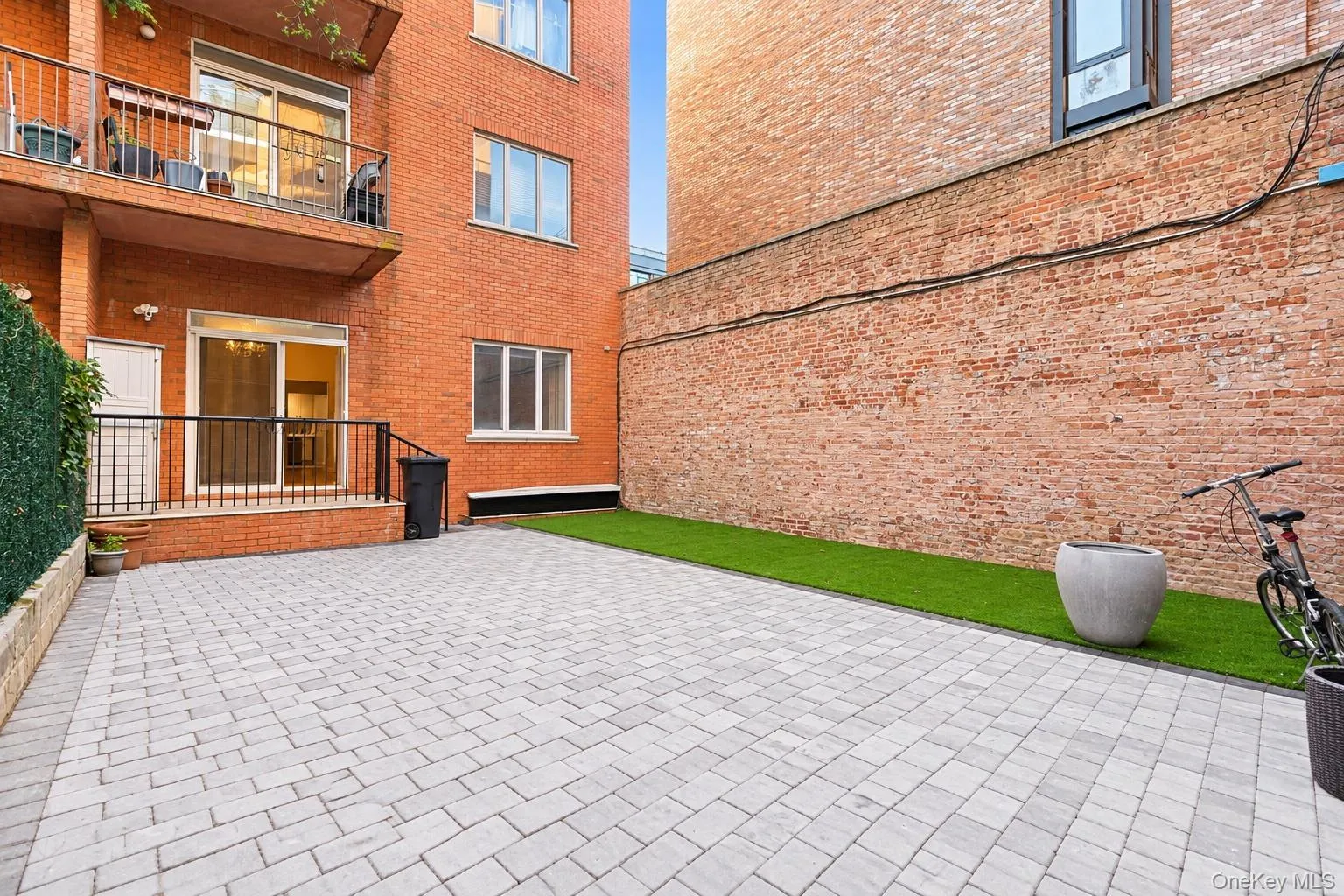 Private outdoor space featuring gray paver hardscaping, a synthetic turf lawn, red brick building exterior, and an elevated metal-railed balcony. Private outdoor space featuring gray paver hardscaping, a synthetic turf lawn, red brick building exterior, and an elevated metal-railed balcony.