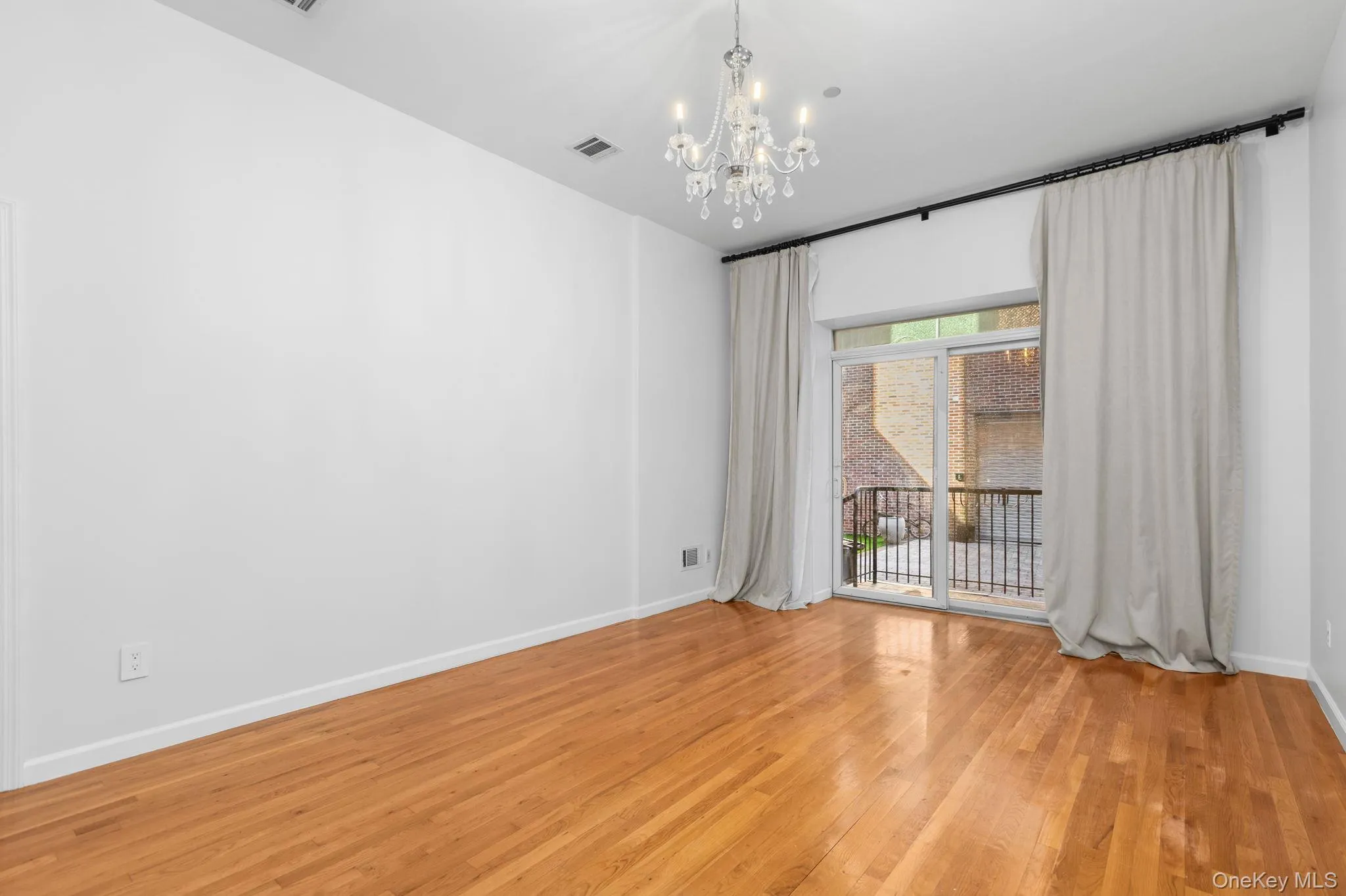 High-ceilinged room featuring wood-finish flooring, a multi-light crystal chandelier, and a sliding glass door to private patio with a decorative black curtain rod High-ceilinged room featuring wood-finish flooring, a multi-light crystal chandelier, and a sliding glass door to private patio with a decorative black curtain rod