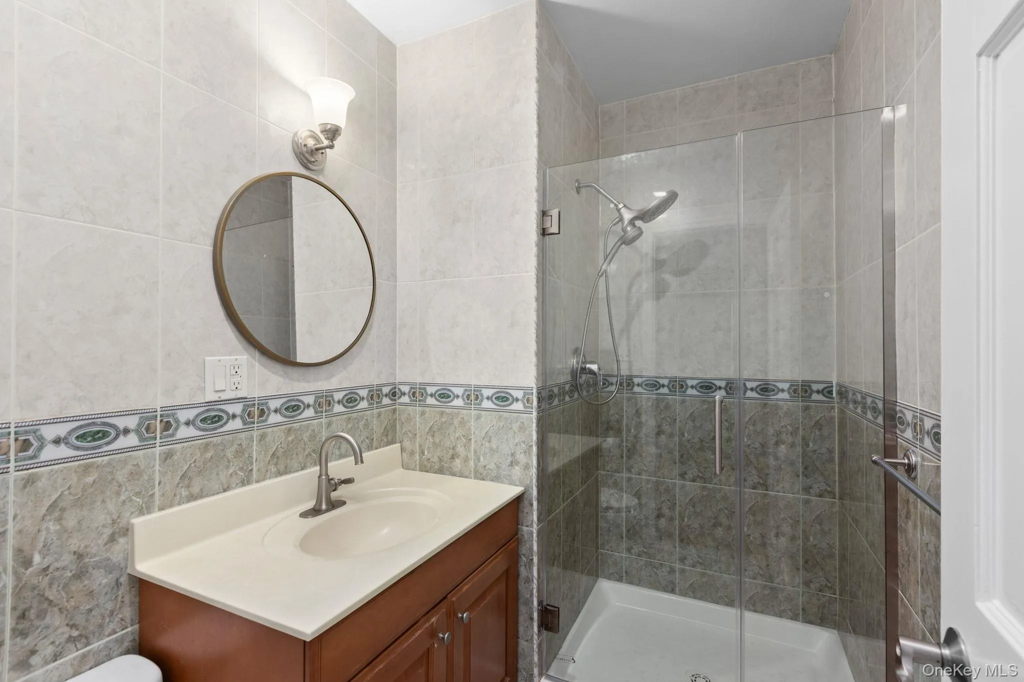 Wood-finish vanity with a molded countertop, glass-enclosed shower featuring a rainfall showerhead, and decorative tiled walls Wood-finish vanity with a molded countertop, glass-enclosed shower featuring a rainfall showerhead, and decorative tiled walls