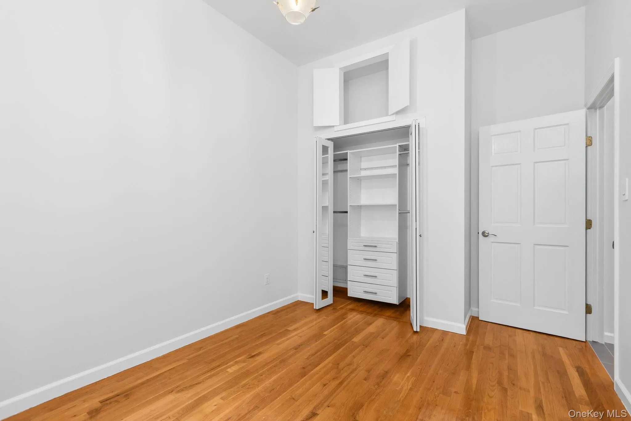 Interior room featuring wood-finish flooring, white walls, and a built-in closet system with integrated shelving and drawers Interior room featuring wood-finish flooring, white walls, and a built-in closet system with integrated shelving and drawers