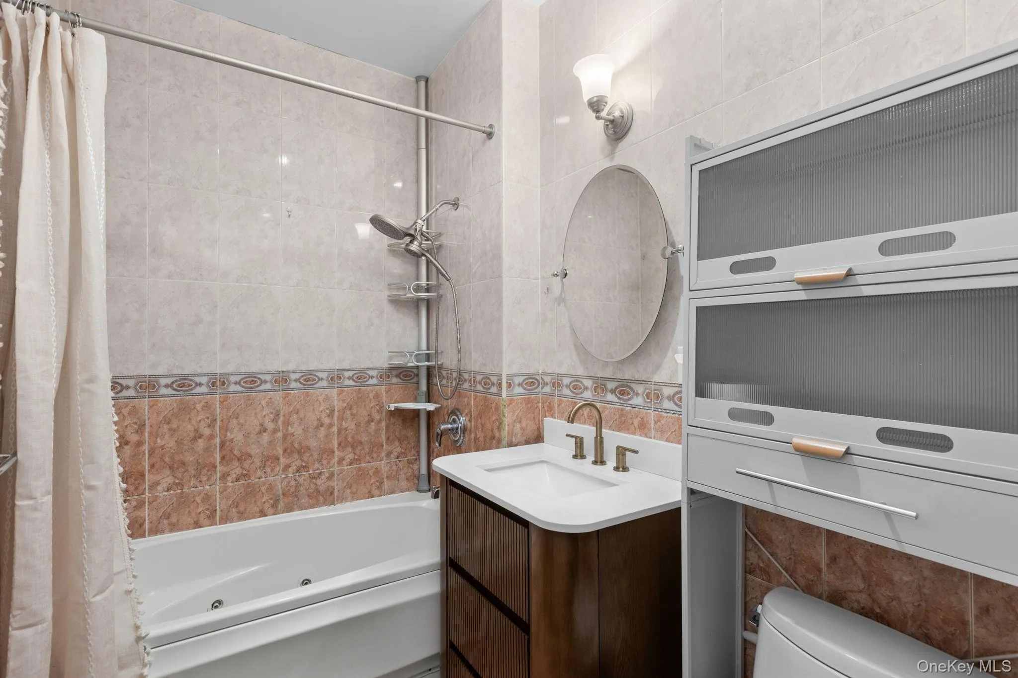 Full-wall tile surround, jetted soaking tub, dark wood-finish vanity with integrated sink, wall-mounted oval mirror, and fixed overhead cabinetry Full-wall tile surround, jetted soaking tub, dark wood-finish vanity with integrated sink, wall-mounted oval mirror, and fixed overhead cabinetry
