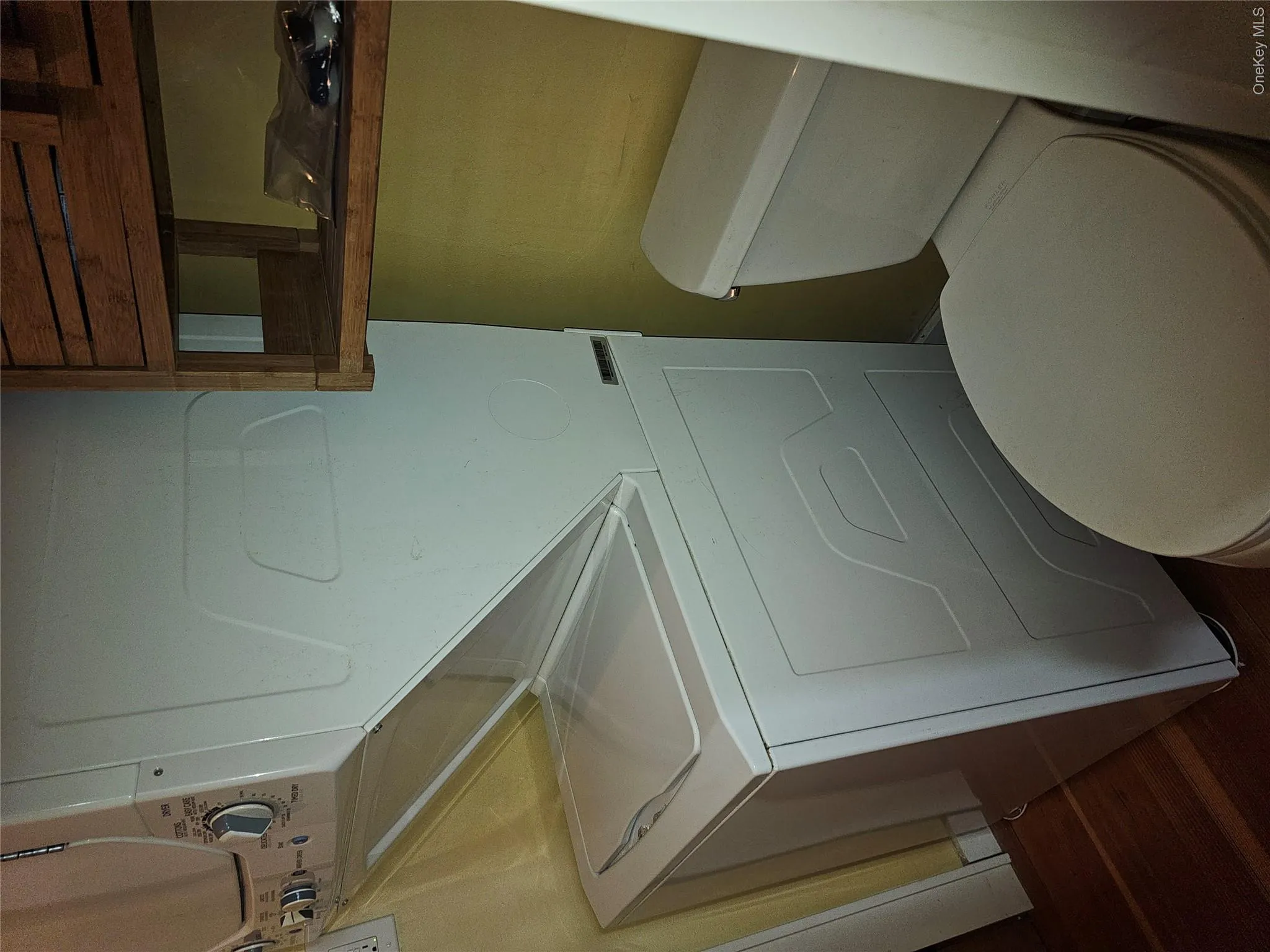 Stacked white laundry center, porcelain toilet, and wall-mounted wooden shelving unit featuring wood-finish flooring Stacked white laundry center, porcelain toilet, and wall-mounted wooden shelving unit featuring wood-finish flooring