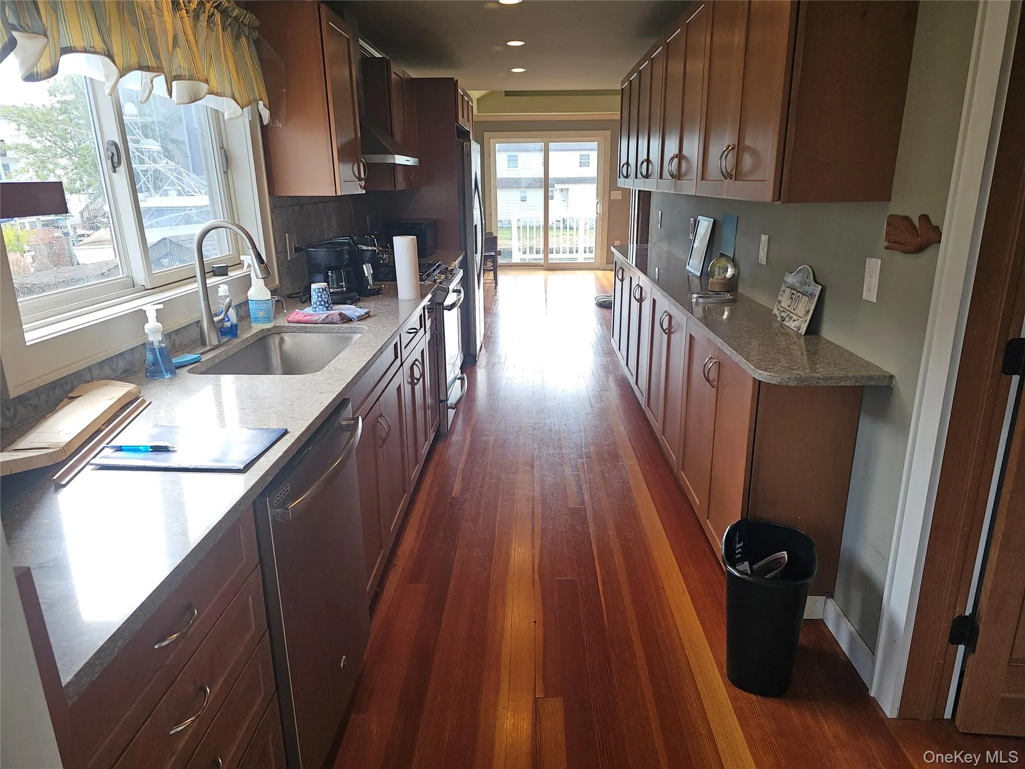 Galley-style kitchen featuring wood-finish flooring, solid-surface countertops, wood cabinetry, stainless steel sink with high-arc faucet, and sliding glass doors Galley-style kitchen featuring wood-finish flooring, solid-surface countertops, wood cabinetry, stainless steel sink with high-arc faucet, and sliding glass doors