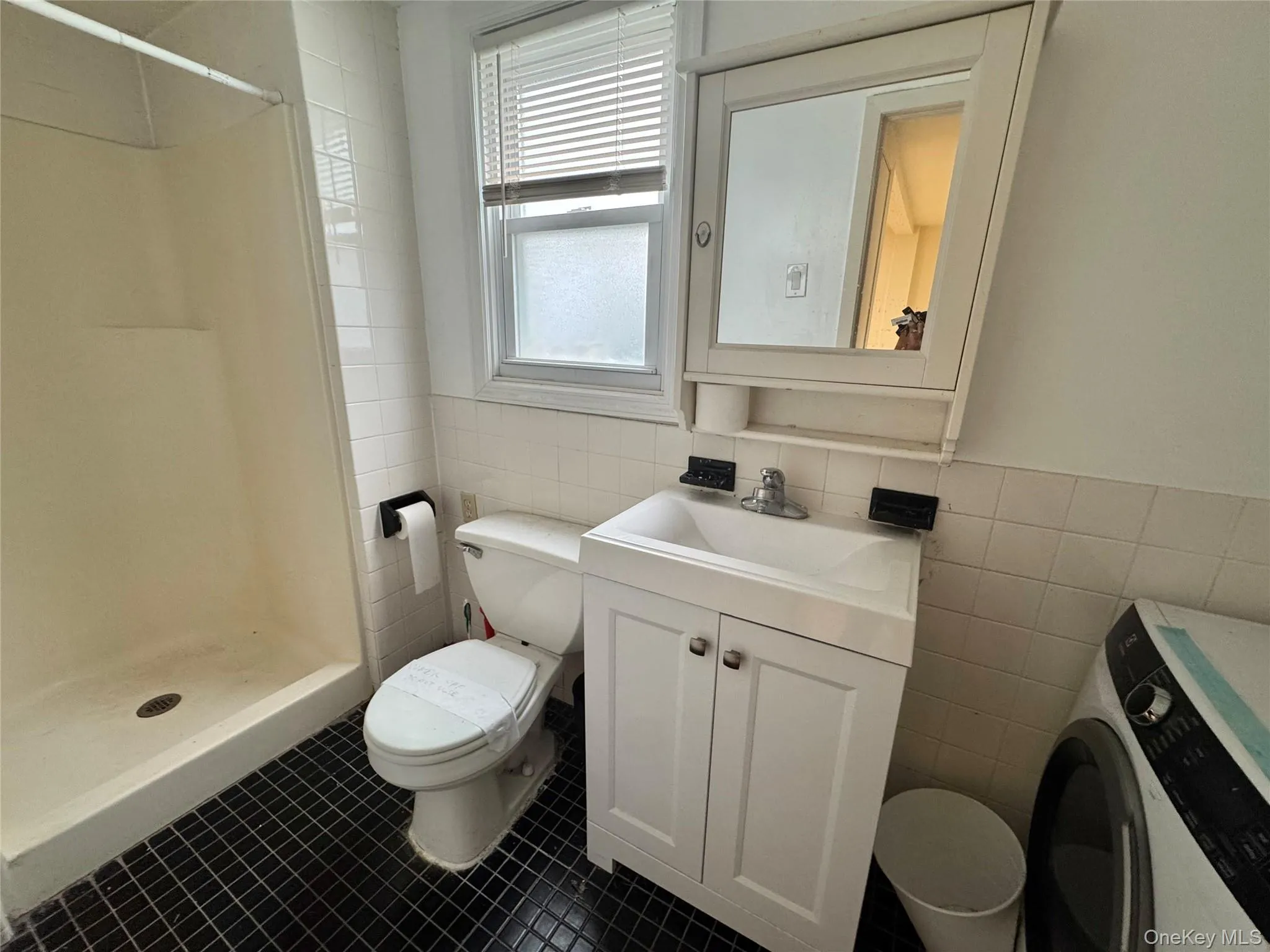 Bathroom featuring white tile wall surround, black square-tile flooring, white vanity with integrated basin, and matching wall-mounted mirrored medicine cabinet Bathroom featuring white tile wall surround, black square-tile flooring, white vanity with integrated basin, and matching wall-mounted mirrored medicine cabinet