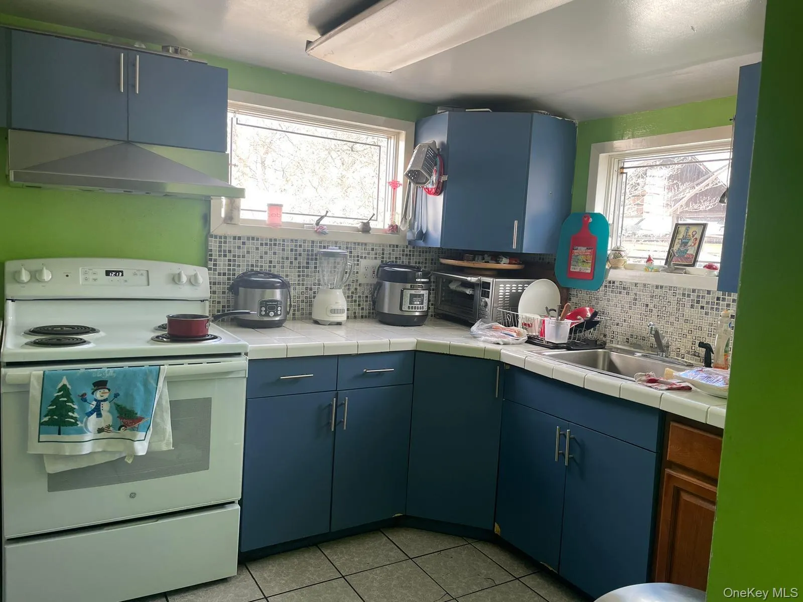 Kitchen featuring white electric range, light tile patterned floors, wall chimney exhaust hood, tasteful backsplash, and a sink Kitchen featuring white electric range, light tile patterned floors, wall chimney exhaust hood, tasteful backsplash, and a sink
