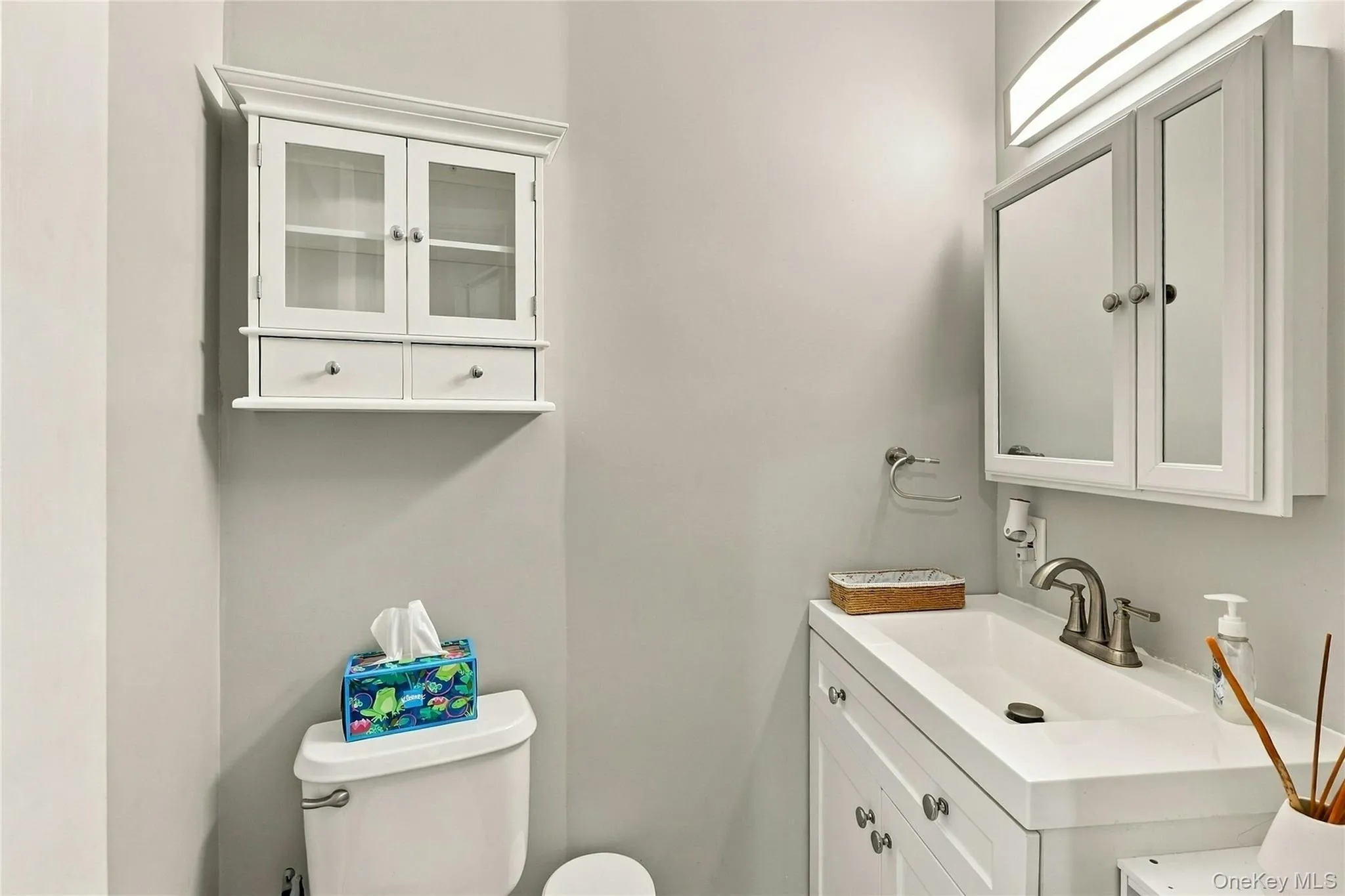 White vanity with integrated rectangular sink and metallic faucet, complemented by a mirrored medicine cabinet, wall-mounted light fixture, and white wall-mounted cabinetry White vanity with integrated rectangular sink and metallic faucet, complemented by a mirrored medicine cabinet, wall-mounted light fixture, and white wall-mounted cabinetry