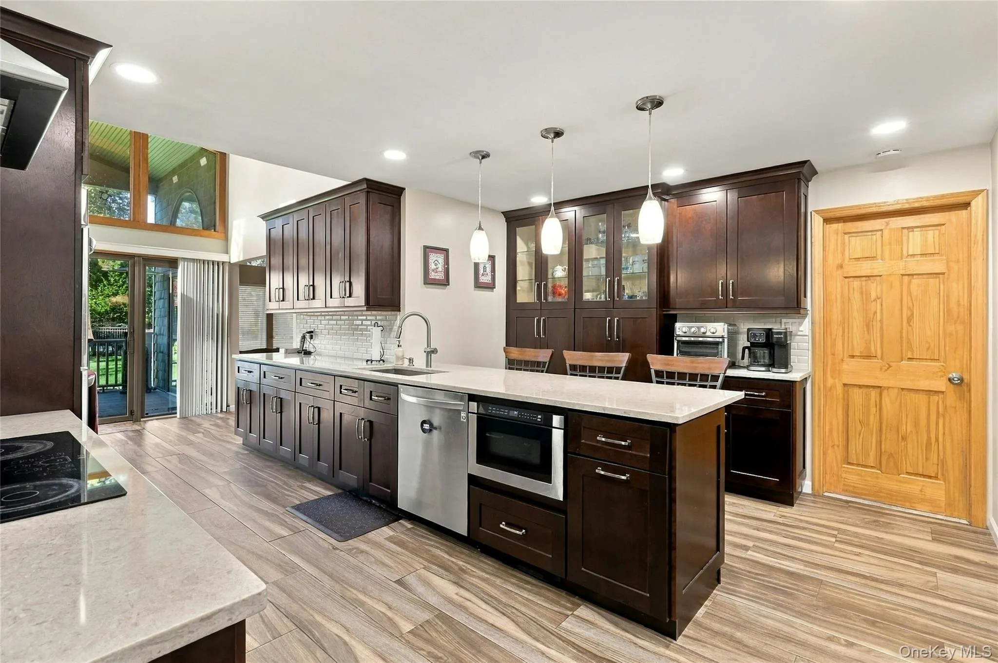 Kitchen featuring dark wood cabinetry, light-toned stone countertops, a central island with integrated microwave, wood-finish flooring, and pendant lighting Kitchen featuring dark wood cabinetry, light-toned stone countertops, a central island with integrated microwave, wood-finish flooring, and pendant lighting