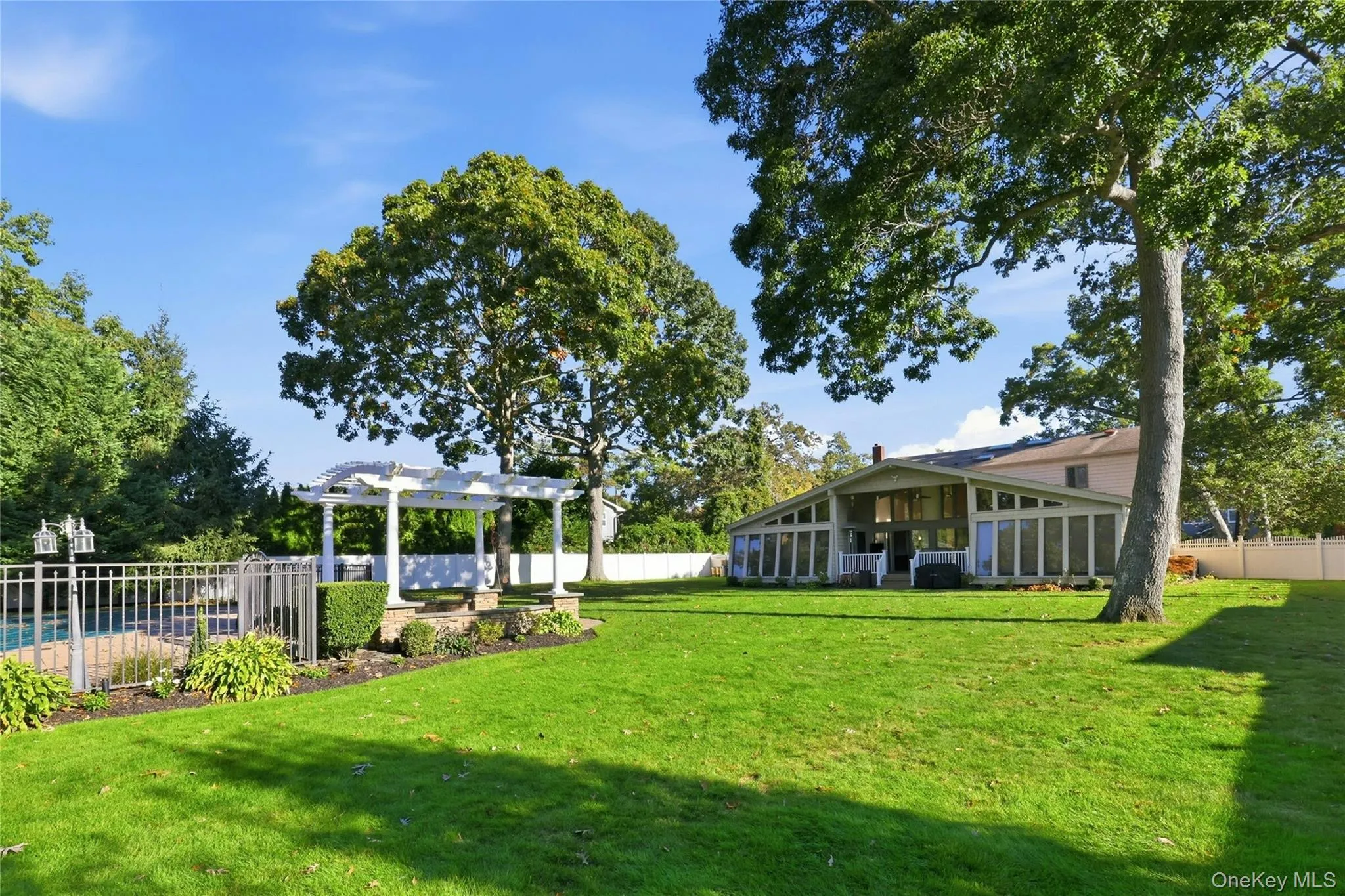 Expansive backyard featuring a sprawling lawn, mature trees, an inground pool with metal perimeter fencing, and a white architectural pergola Expansive backyard featuring a sprawling lawn, mature trees, an inground pool with metal perimeter fencing, and a white architectural pergola
