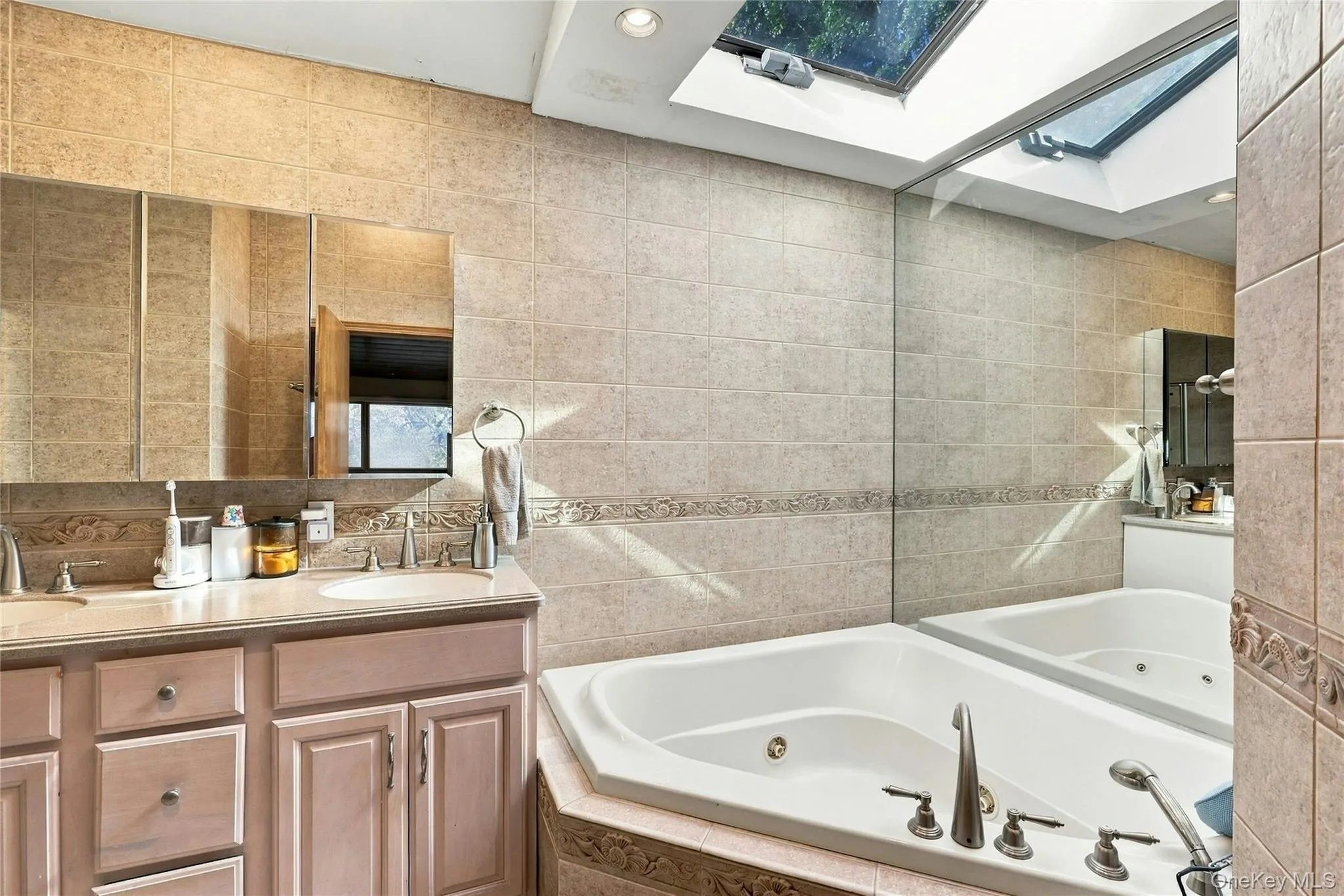 Custom wooden vanity with dual recessed-mirror cabinets, corner jetted tub, wall-to-wall tiling with decorative trim, and an expansive overhead skylight Custom wooden vanity with dual recessed-mirror cabinets, corner jetted tub, wall-to-wall tiling with decorative trim, and an expansive overhead skylight