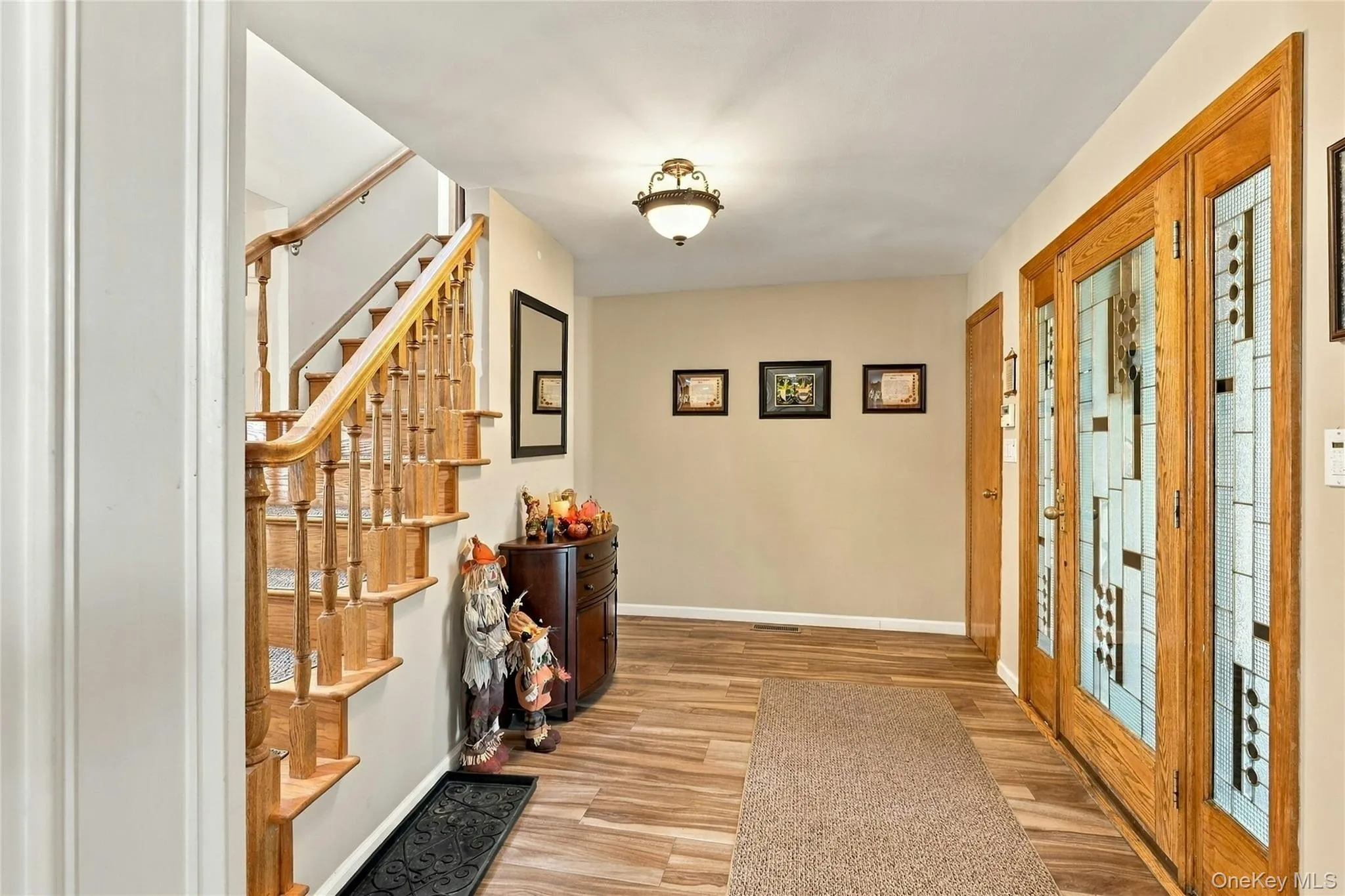 Entry foyer featuring a wood-finish staircase with a decorative railing, double-door entrance with geometric glass panels, wood-finish flooring, and a flush-mount light fixture Entry foyer featuring a wood-finish staircase with a decorative railing, double-door entrance with geometric glass panels, wood-finish flooring, and a flush-mount light fixture