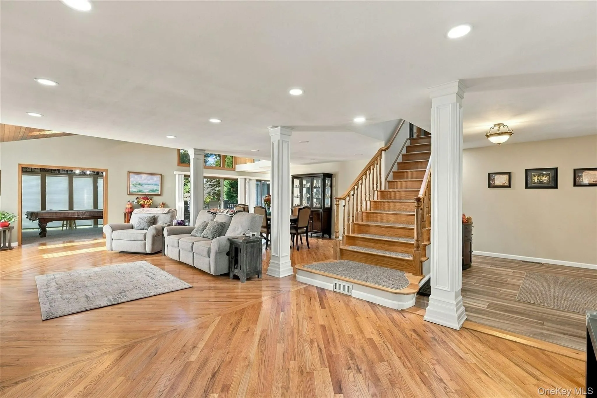 Open-concept interior featuring wood-finish flooring, recessed lighting, decorative white columns, and a wooden staircase with a matching handrail Open-concept interior featuring wood-finish flooring, recessed lighting, decorative white columns, and a wooden staircase with a matching handrail