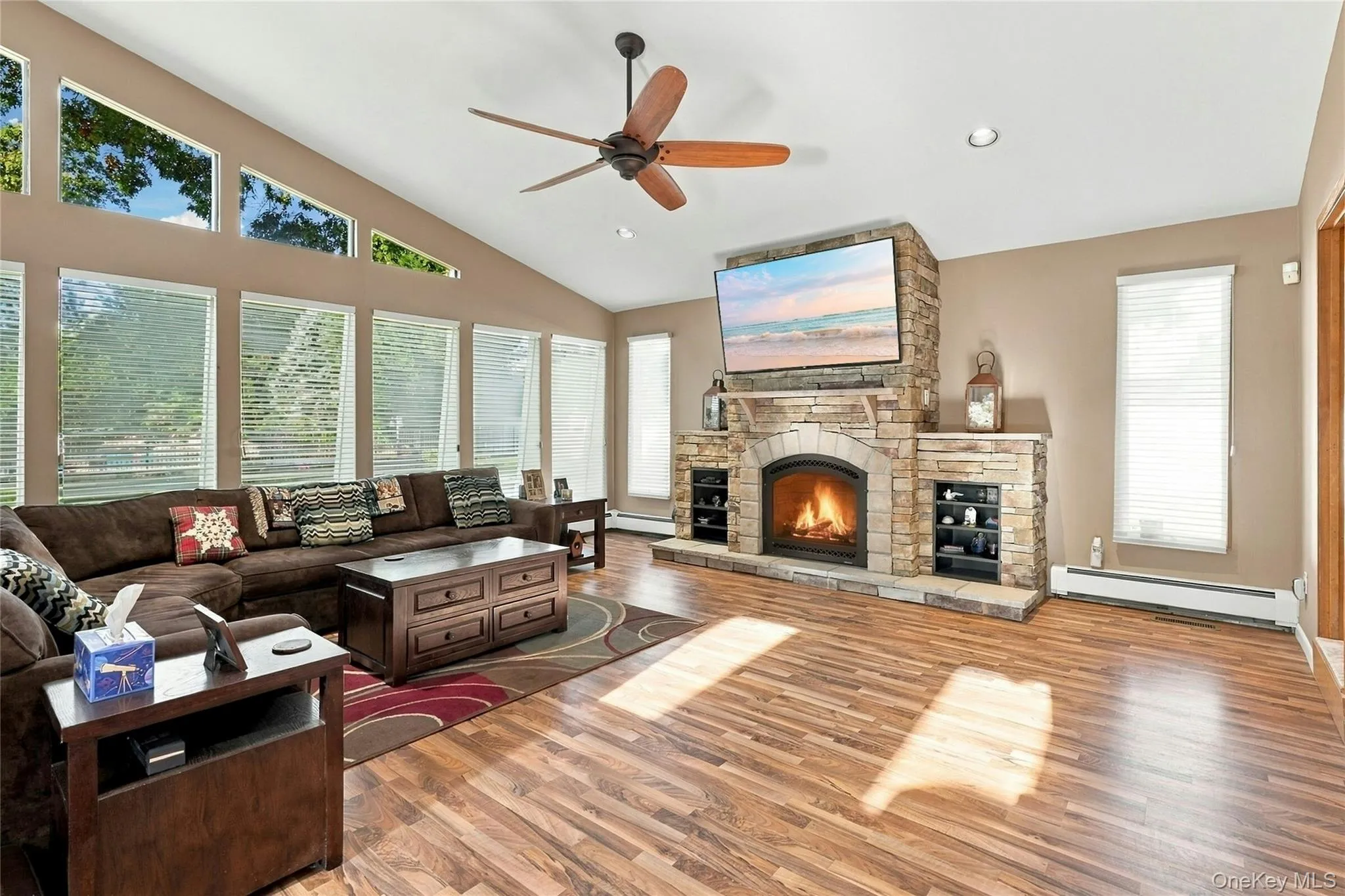 Living room featuring vaulted ceilings, floor-to-ceiling stone-faced fireplace with hearth, wood-finish flooring, multi-pane window configuration, and a wooden ceiling fan Living room featuring vaulted ceilings, floor-to-ceiling stone-faced fireplace with hearth, wood-finish flooring, multi-pane window configuration, and a wooden ceiling fan