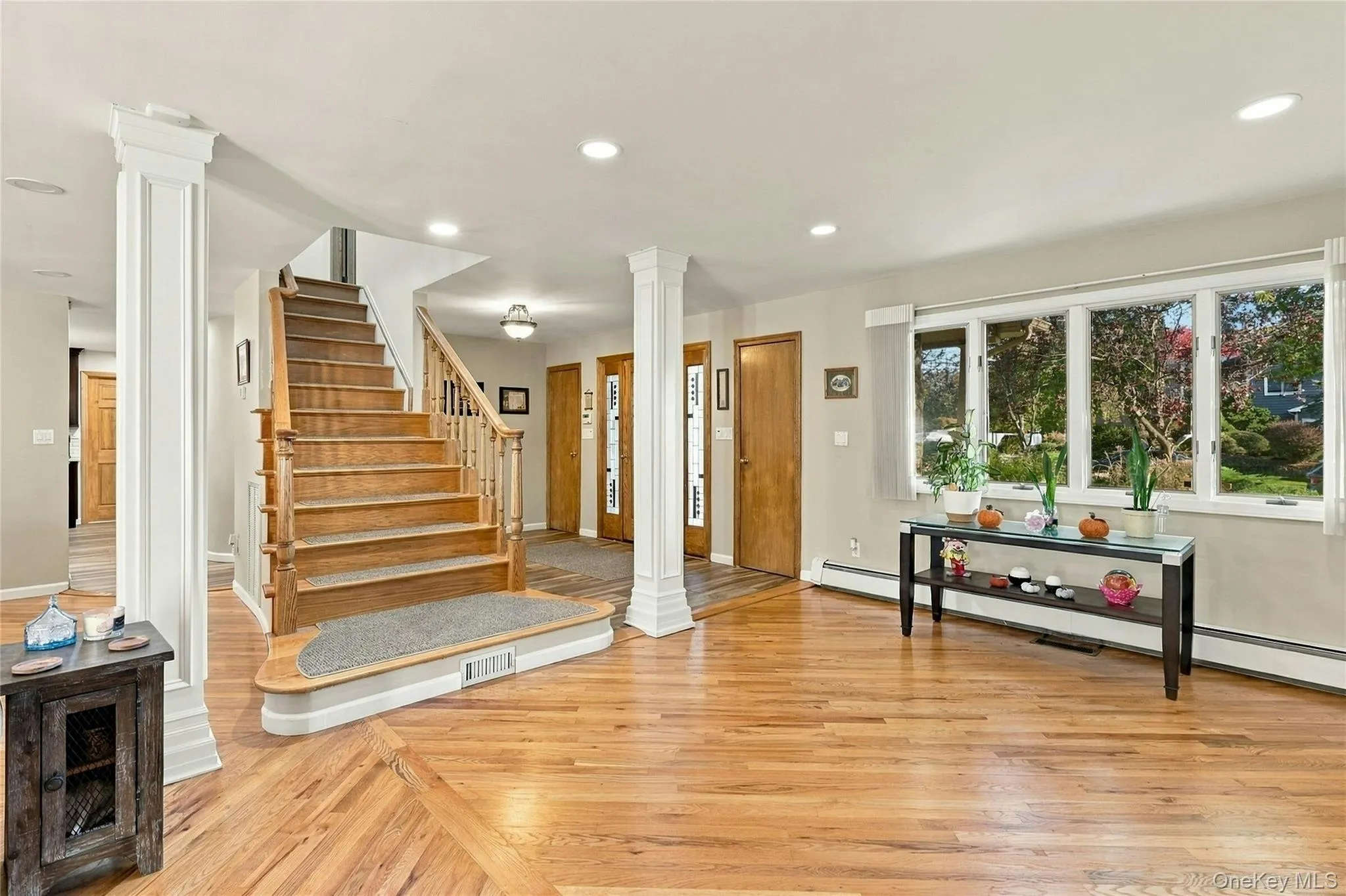 Open-concept interior featuring wood-finish flooring, a central staircase with a wood-finish handrail, architectural columns, multiple wood-finish interior doors, and a large multi-pane window Open-concept interior featuring wood-finish flooring, a central staircase with a wood-finish handrail, architectural columns, multiple wood-finish interior doors, and a large multi-pane window