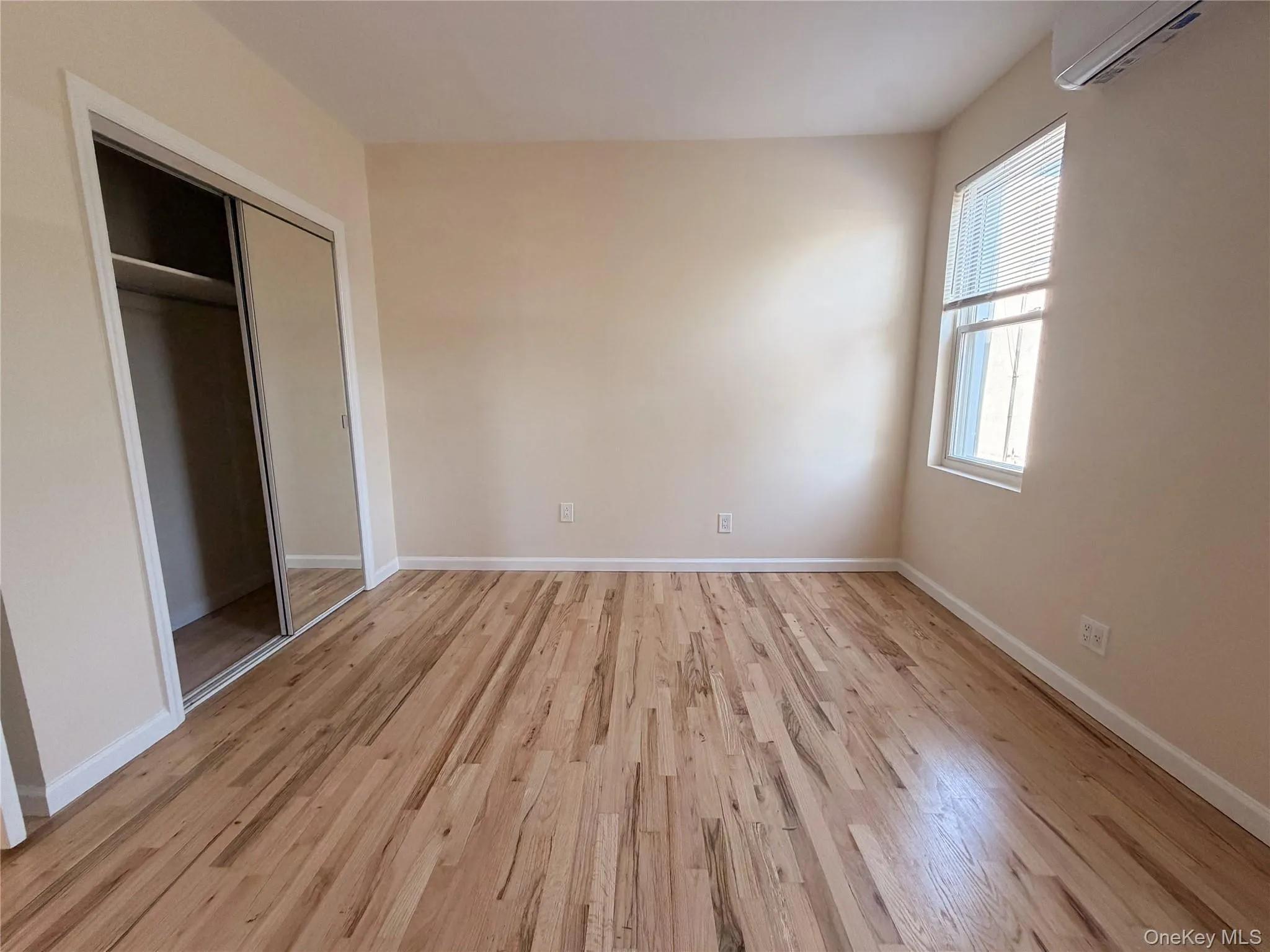 Room featuring wood-finish flooring, sliding mirror-panel closet doors, and a wall-mounted climate control unit. Room featuring wood-finish flooring, sliding mirror-panel closet doors, and a wall-mounted climate control unit.