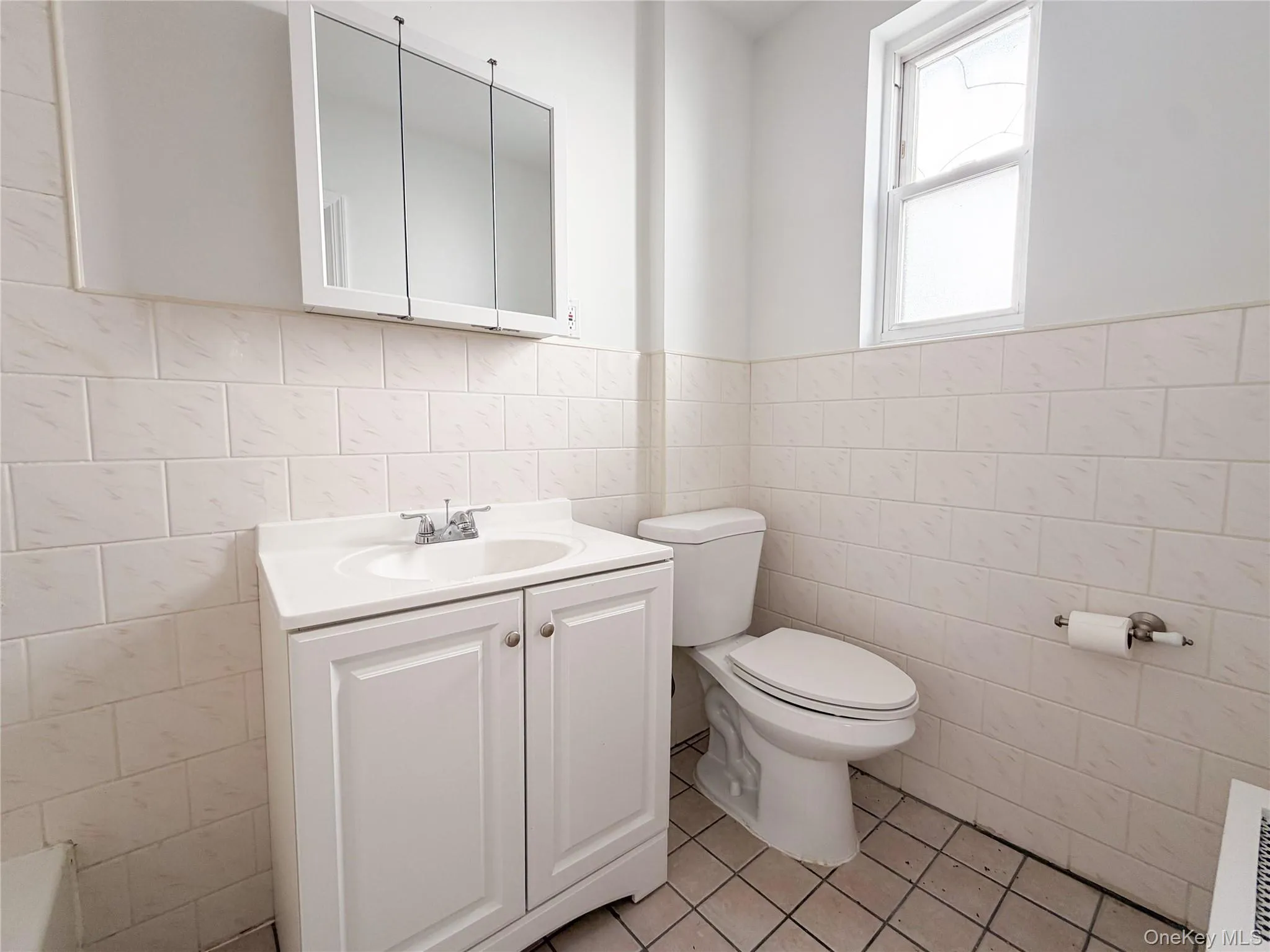 Bathroom featuring a vanity with an integrated basin, a three-panel medicine cabinet, tiled wall surfaces, and a window. Bathroom featuring a vanity with an integrated basin, a three-panel medicine cabinet, tiled wall surfaces, and a window.