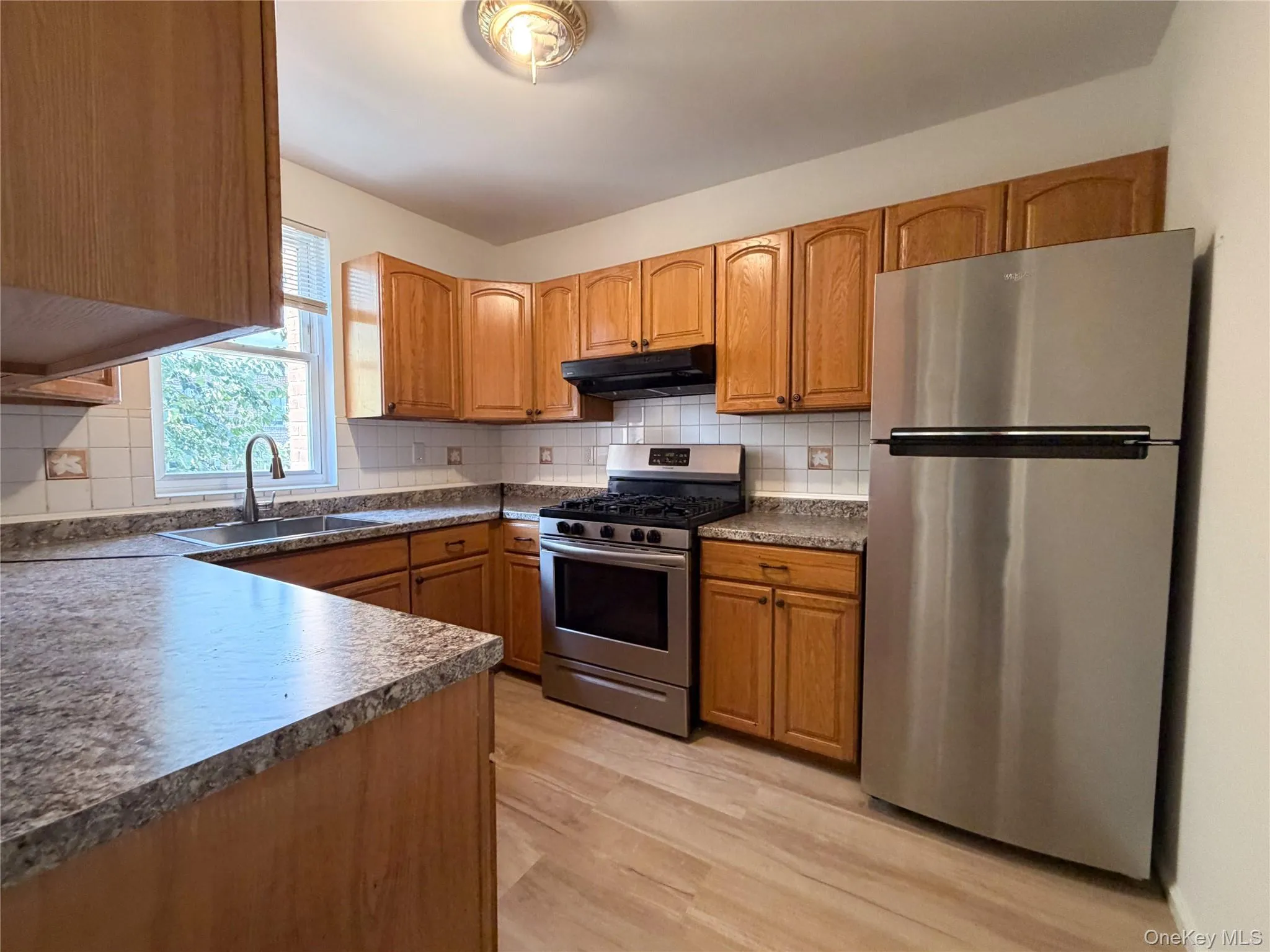 Kitchen featuring wood cabinetry, stone-finish countertops, tiled backsplash, stainless steel appliances, and wood-finish flooring. Kitchen featuring wood cabinetry, stone-finish countertops, tiled backsplash, stainless steel appliances, and wood-finish flooring.