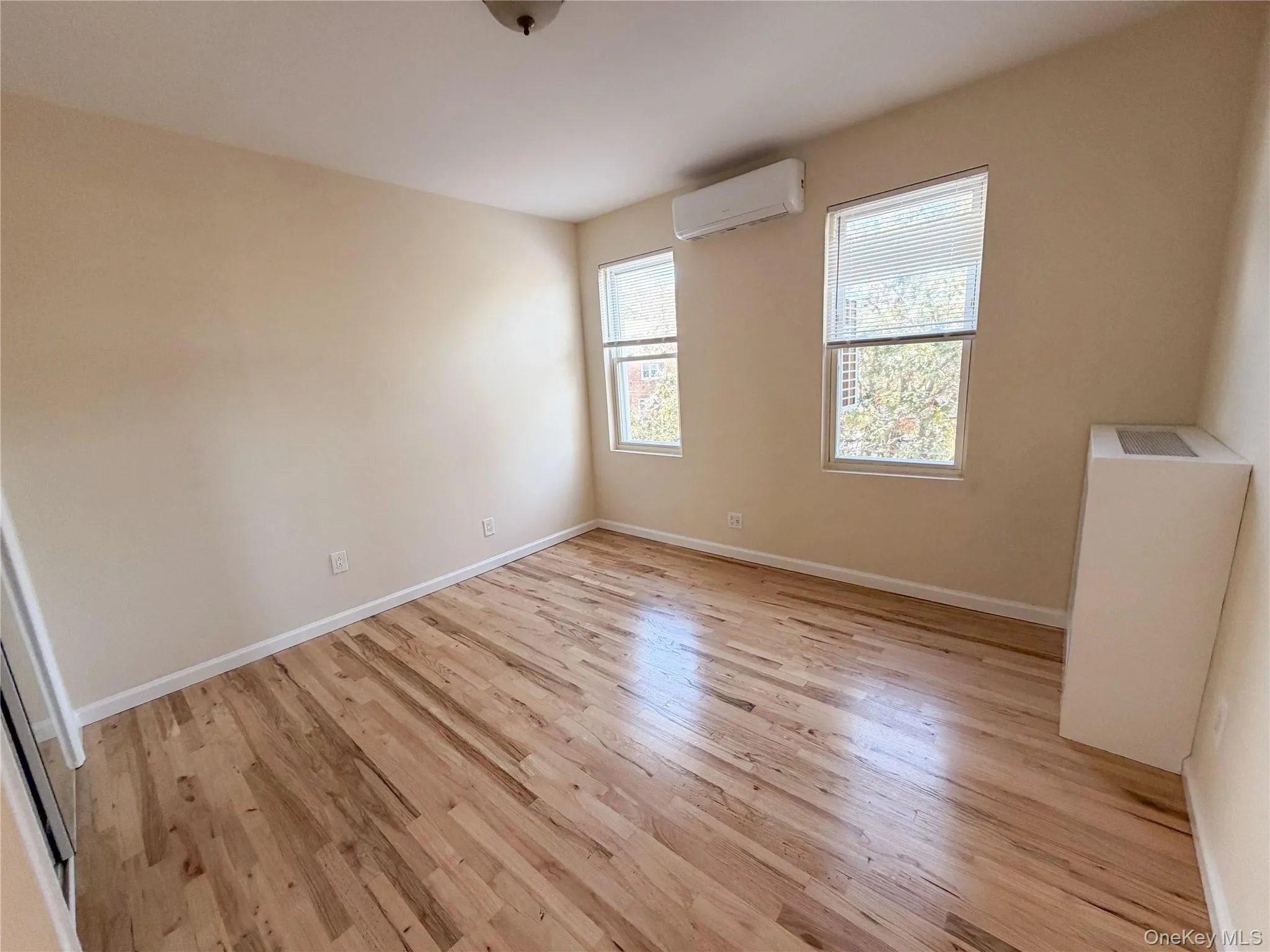 Interior room featuring wood-finish flooring, two double-hung windows, and a wall-mounted ductless heating and cooling unit. Interior room featuring wood-finish flooring, two double-hung windows, and a wall-mounted ductless heating and cooling unit.