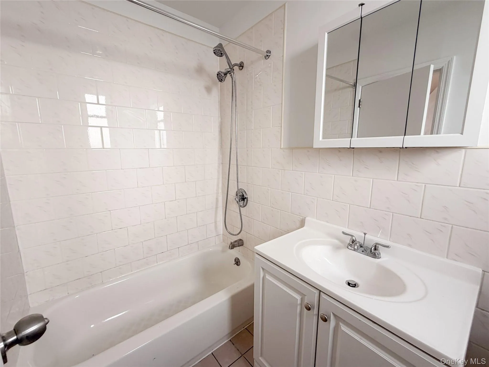 Bathroom featuring a white vanity cabinet with an integrated sink, tri-fold mirrored medicine cabinet, and a shower-tub combination surrounded by light-toned rectangular wall tile. Bathroom featuring a white vanity cabinet with an integrated sink, tri-fold mirrored medicine cabinet, and a shower-tub combination surrounded by light-toned rectangular wall tile.