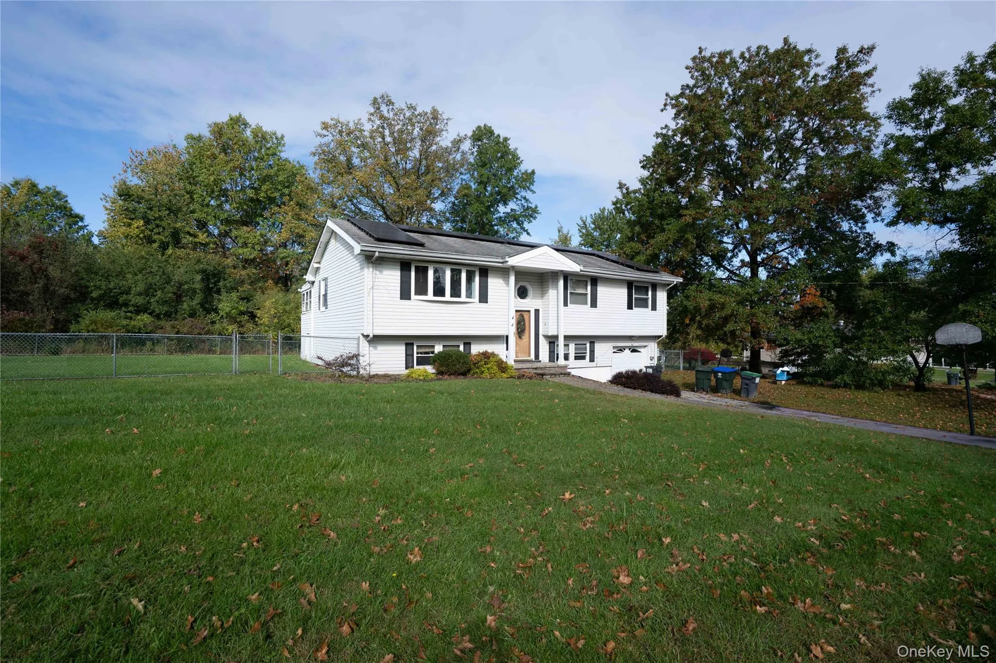 Split-level exterior featuring light-colored siding, black shutter accents, solar panels, and a partially fenced yard with mature trees Split-level exterior featuring light-colored siding, black shutter accents, solar panels, and a partially fenced yard with mature trees