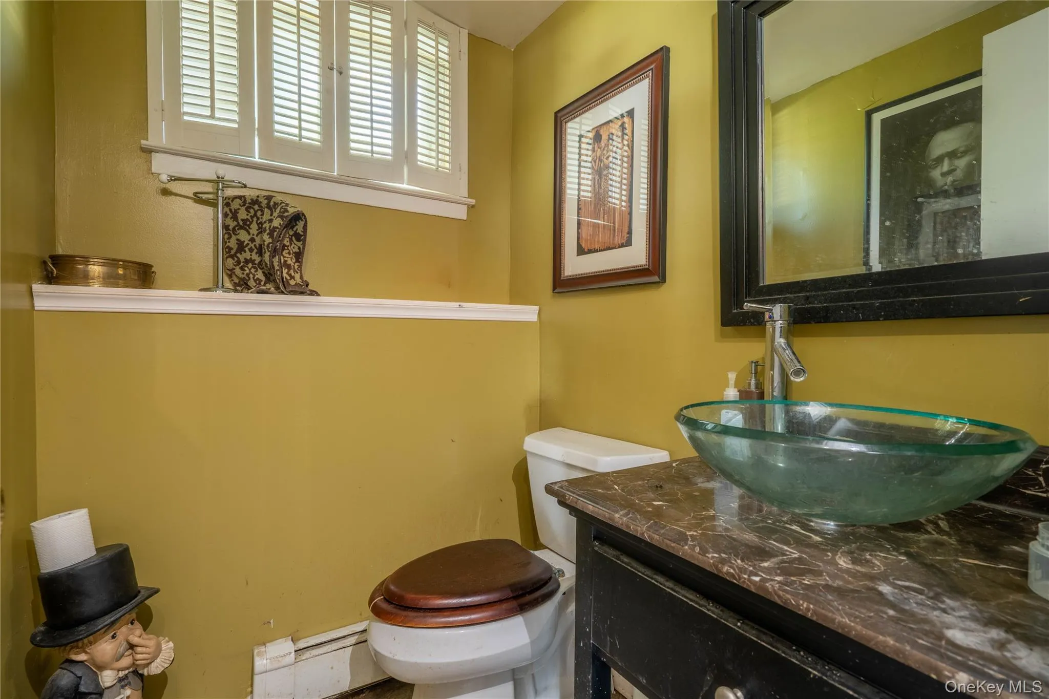 Restroom featuring a glass vessel sink, stone-finish countertop, dark wood-finish vanity, louvered shutters, and framed mirror Restroom featuring a glass vessel sink, stone-finish countertop, dark wood-finish vanity, louvered shutters, and framed mirror