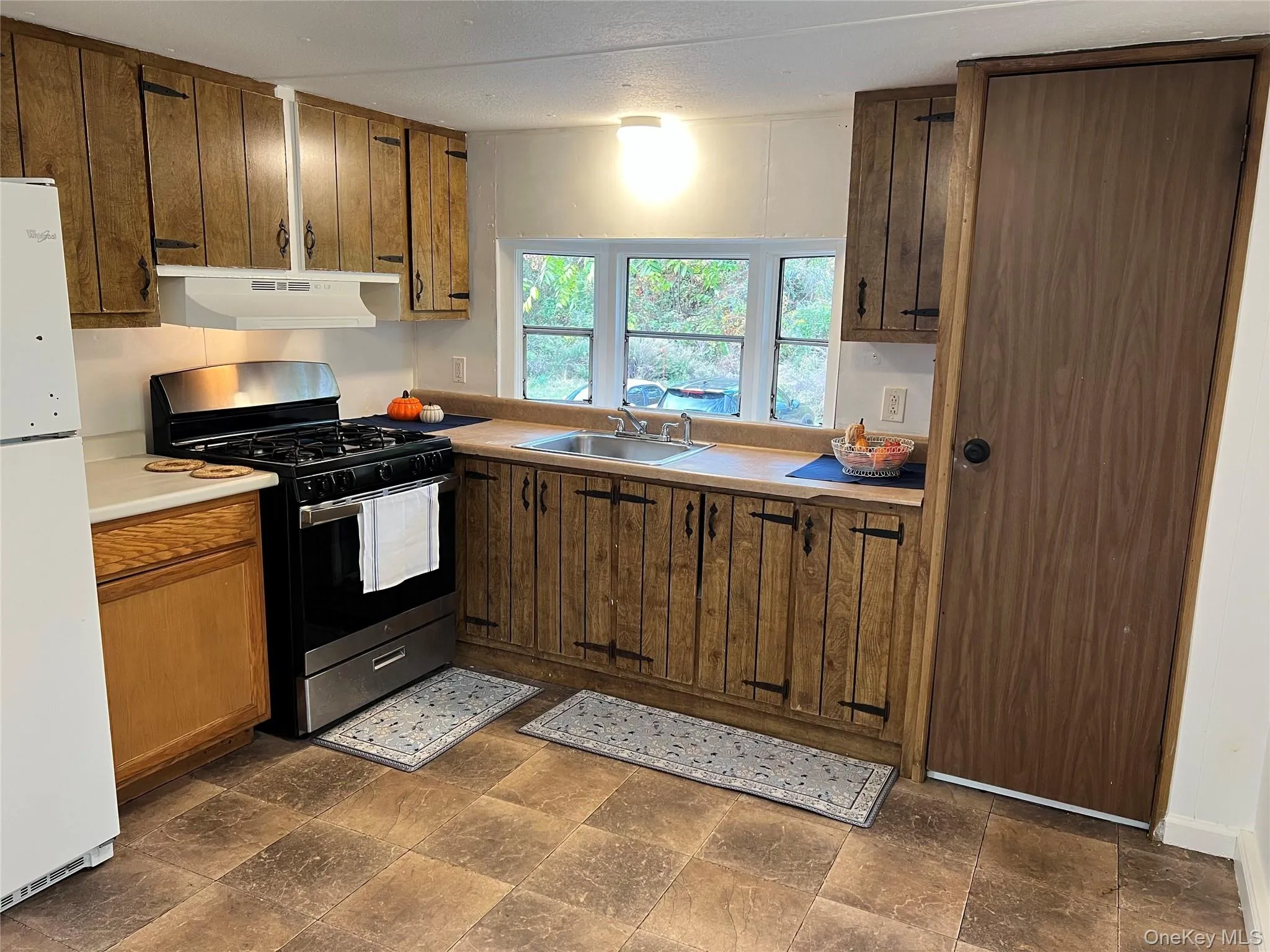 Kitchen with stainless steel gas range, freestanding refrigerator, light countertops, under cabinet range hood, and dark stone finish floors Kitchen with stainless steel gas range, freestanding refrigerator, light countertops, under cabinet range hood, and dark stone finish floors