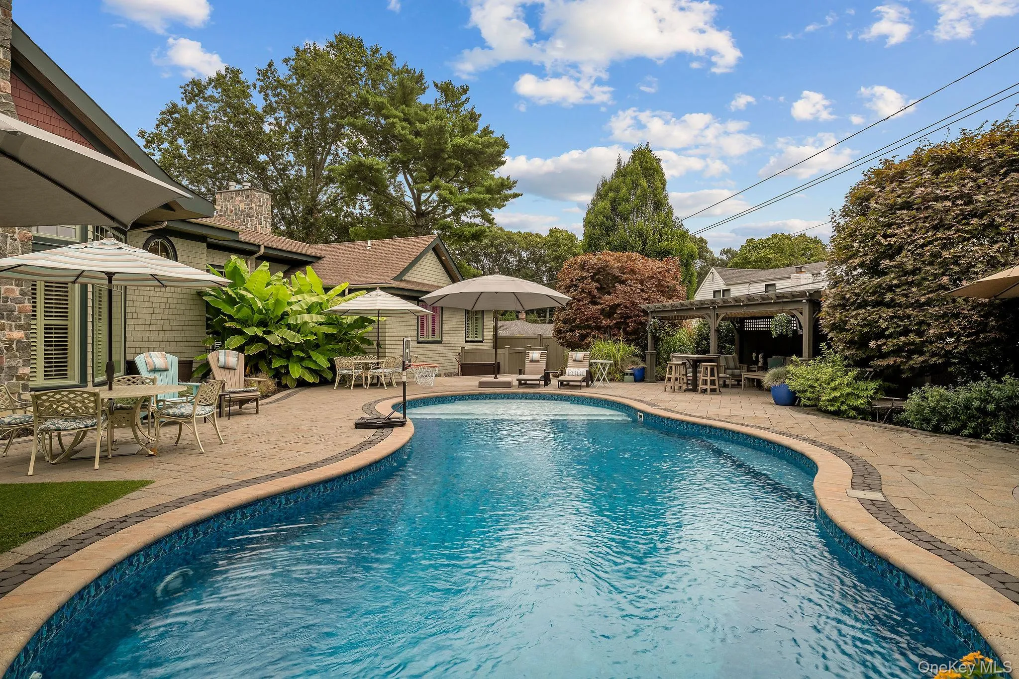 Curved in-ground swimming pool featuring stone-paver decking, a built-in pergola, and stone masonry exterior siding Curved in-ground swimming pool featuring stone-paver decking, a built-in pergola, and stone masonry exterior siding