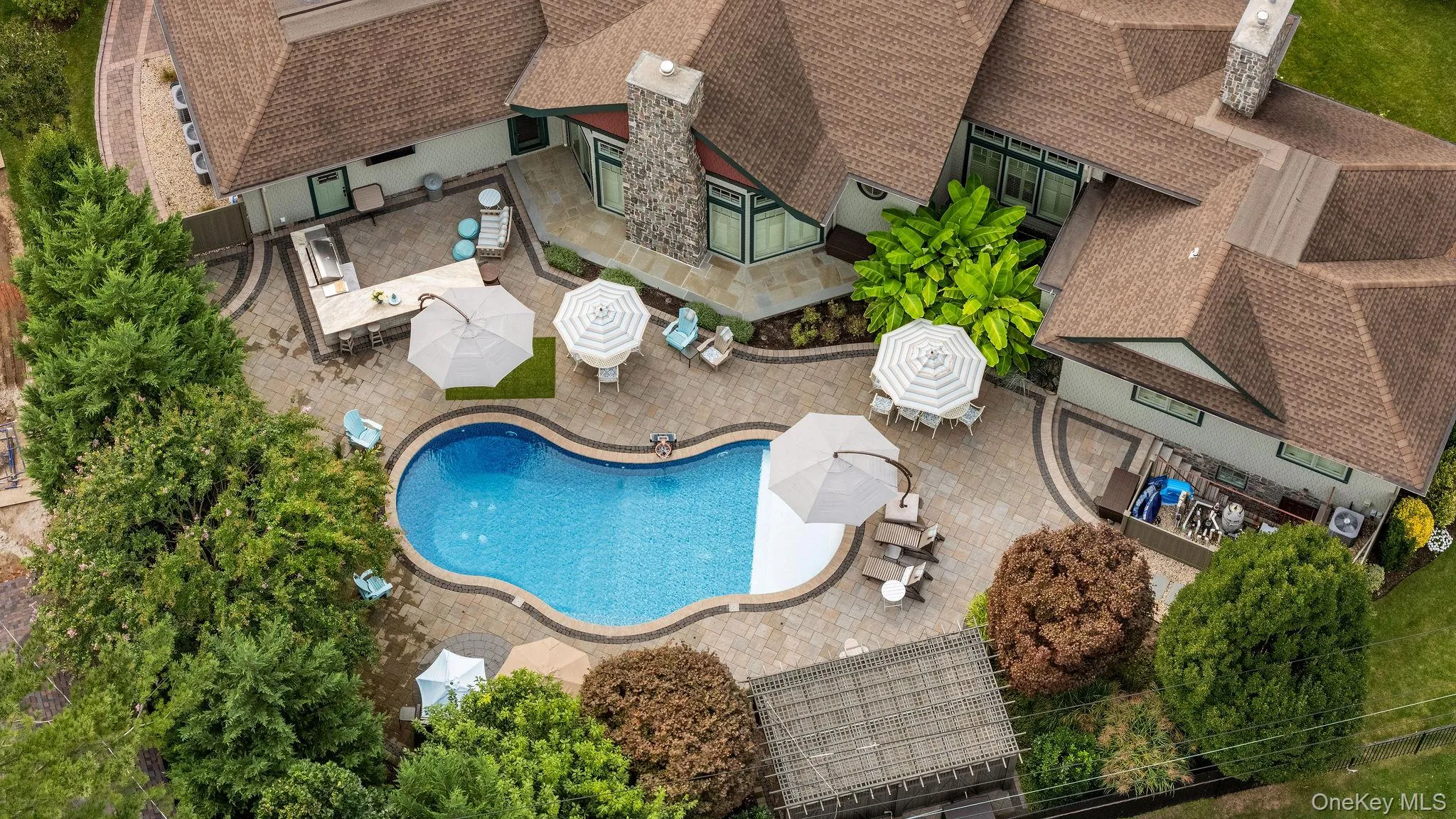 Aerial perspective of a sprawling residence featuring a custom-shaped swimming pool, extensive stone paver hardscaping, a built-in outdoor kitchen with countertop, and two stone masonry chimneys Aerial perspective of a sprawling residence featuring a custom-shaped swimming pool, extensive stone paver hardscaping, a built-in outdoor kitchen with countertop, and two stone masonry chimneys