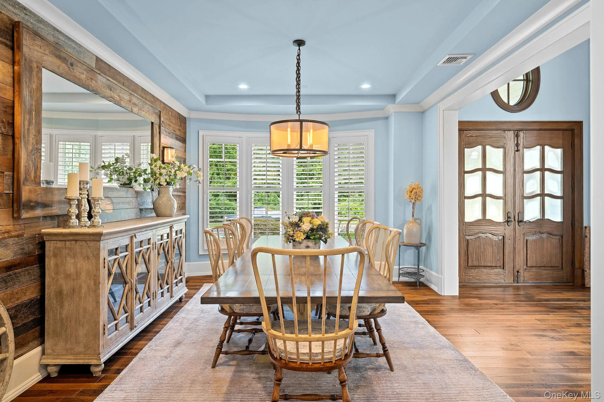 Dining area featuring wood-finish plank flooring, wood-paneled accent wall, tray ceiling with recessed lighting, bay windows with plantation shutters, and a central chandelier Dining area featuring wood-finish plank flooring, wood-paneled accent wall, tray ceiling with recessed lighting, bay windows with plantation shutters, and a central chandelier