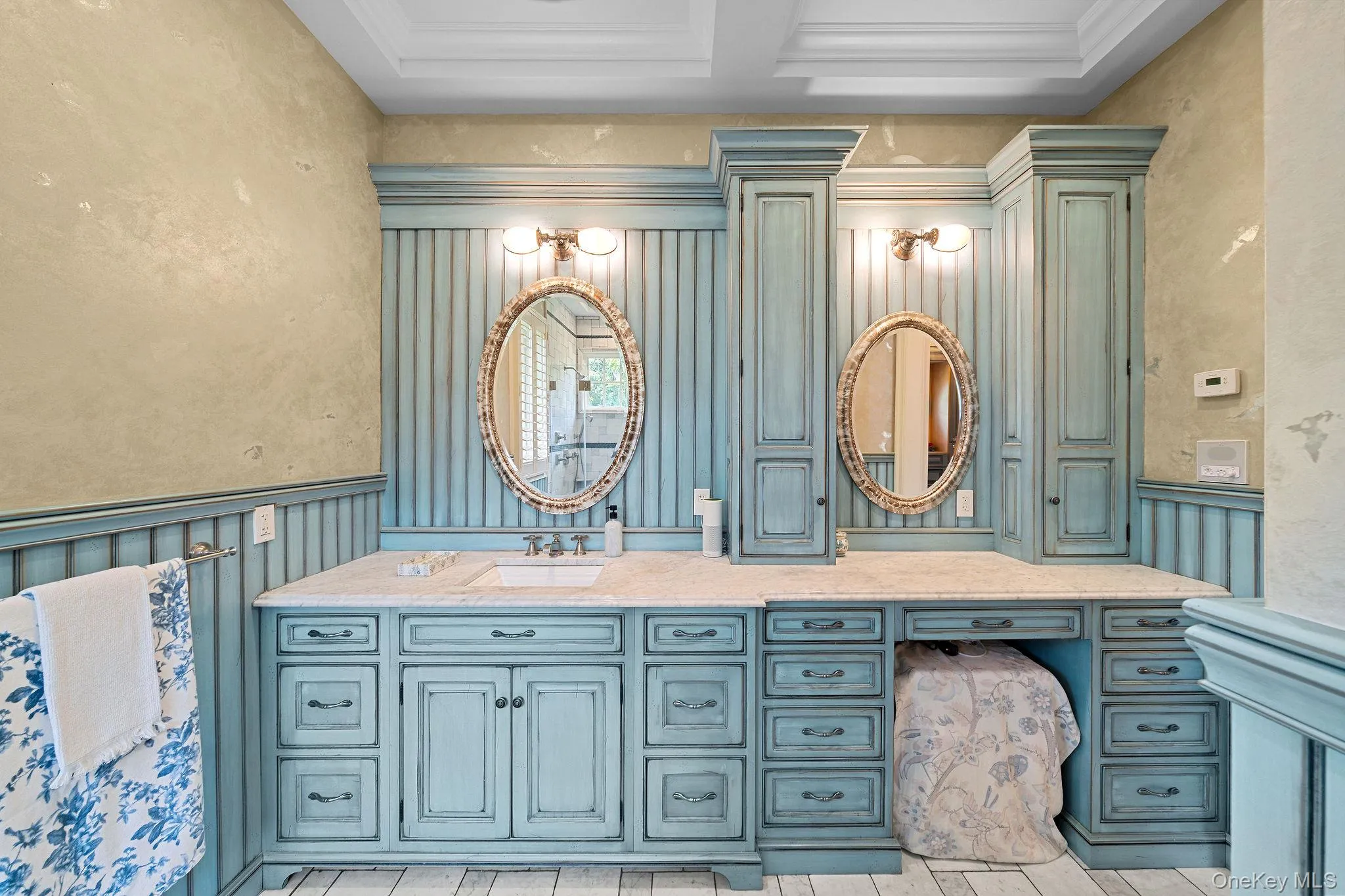 Dual-sink vanity featuring custom blue cabinetry, stone countertop, integrated floor-to-ceiling storage towers, beadboard wainscoting, and oval wall mirrors Dual-sink vanity featuring custom blue cabinetry, stone countertop, integrated floor-to-ceiling storage towers, beadboard wainscoting, and oval wall mirrors