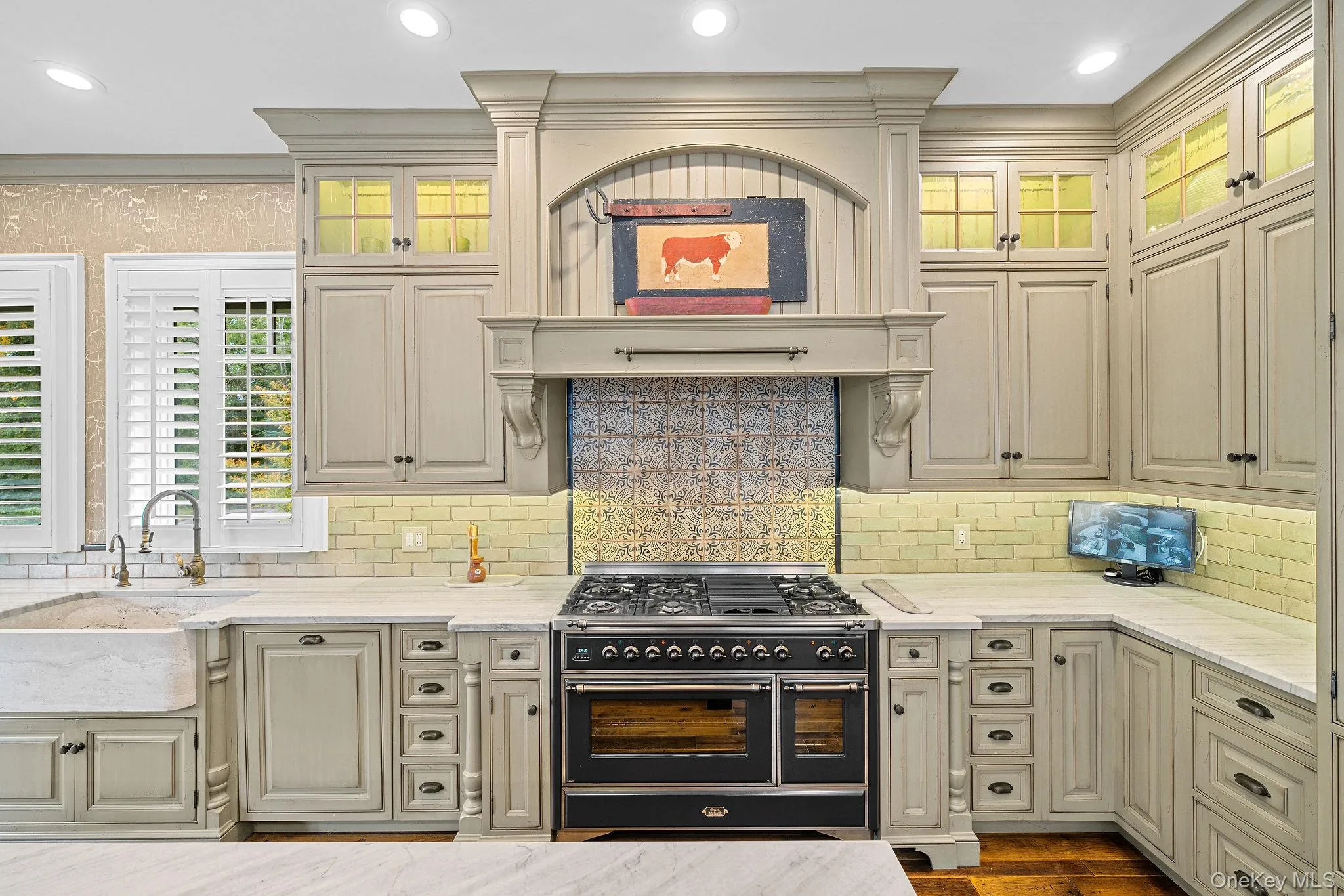 Custom-painted cabinetry featuring glass-front upper displays and a decorative hood enclosure, professional-grade dual-fuel range, farmhouse sink with gooseneck faucet, patterned tile backsplash, and stone countertops Custom-painted cabinetry featuring glass-front upper displays and a decorative hood enclosure, professional-grade dual-fuel range, farmhouse sink with gooseneck faucet, patterned tile backsplash, and stone countertops